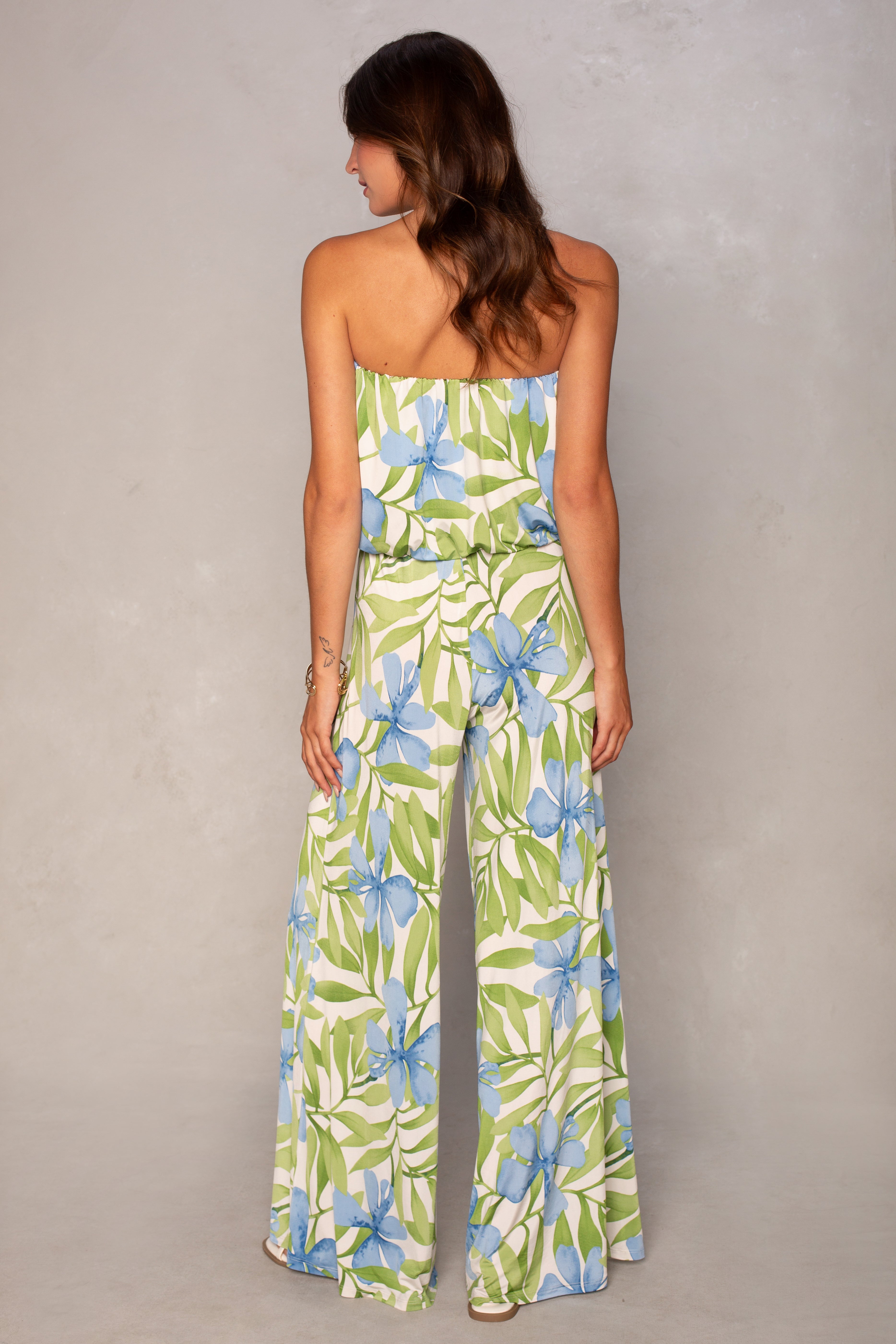 A woman with loose brown waves stands with her back to the camera in the Ari Strapless Easy Jumpsuit by Isla, featuring a tropical print of green leaves and blue flowers and wide leg pants.