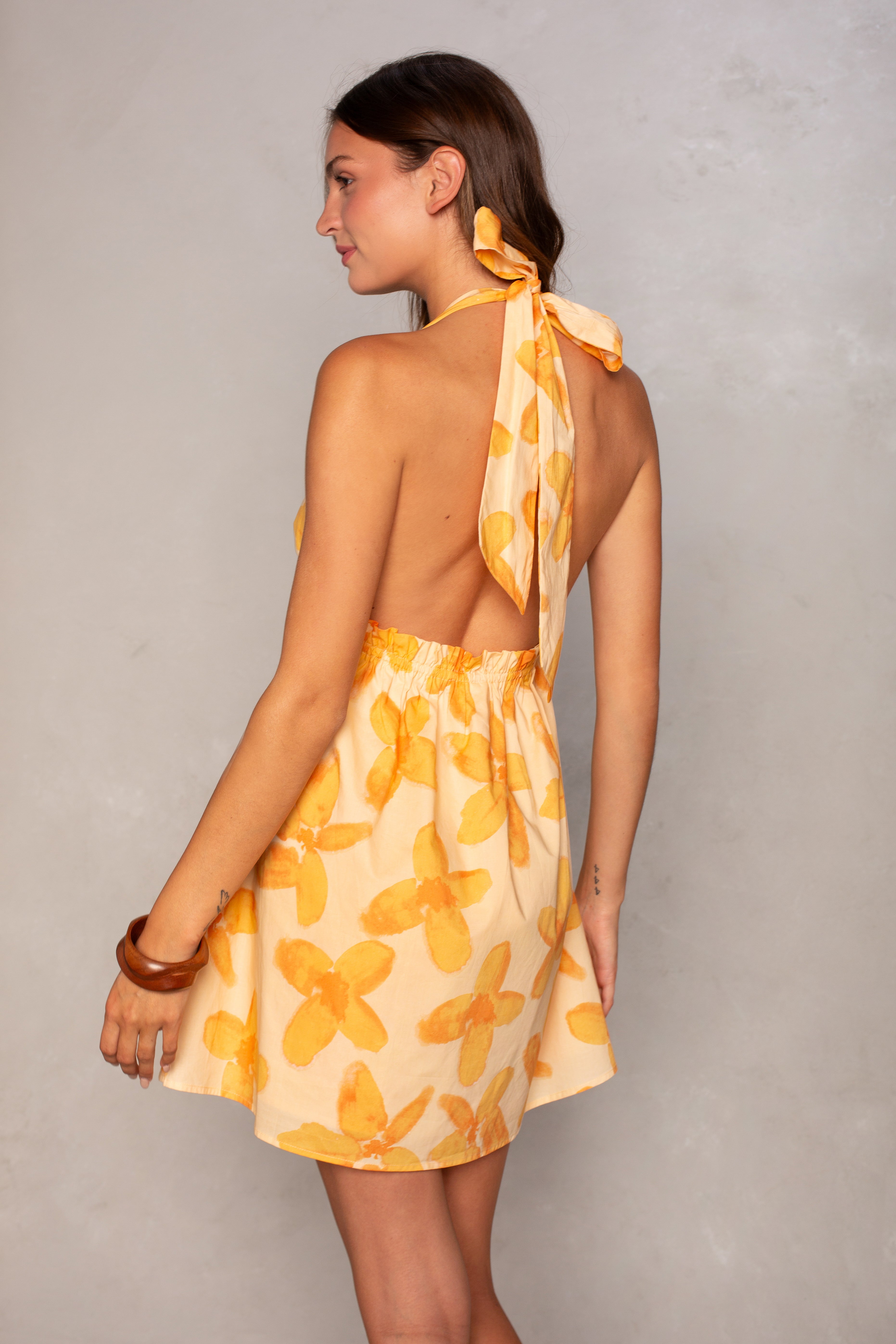 A woman with light brown hair stands against a neutral wall, wearing the Isla Mira Halter Mini Dress—a yellow floral print dress featuring a halter neckline and open back bow tie detail—paired with a brown bracelet.