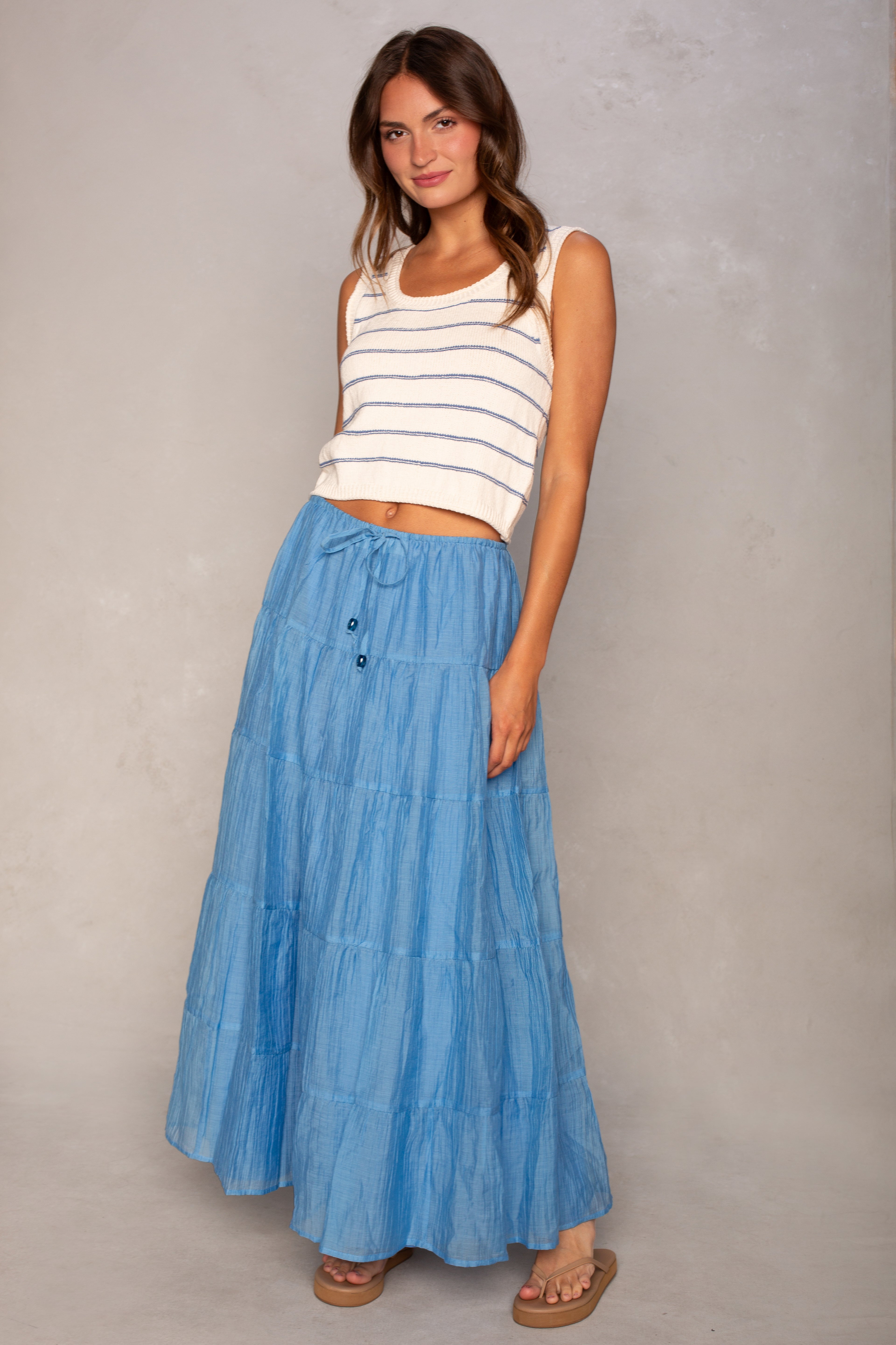 A woman with wavy brown hair stands against a neutral background wearing the Isla Piper Sleeveless Sweater, a long tiered blue skirt, and tan sandals. She smiles slightly.