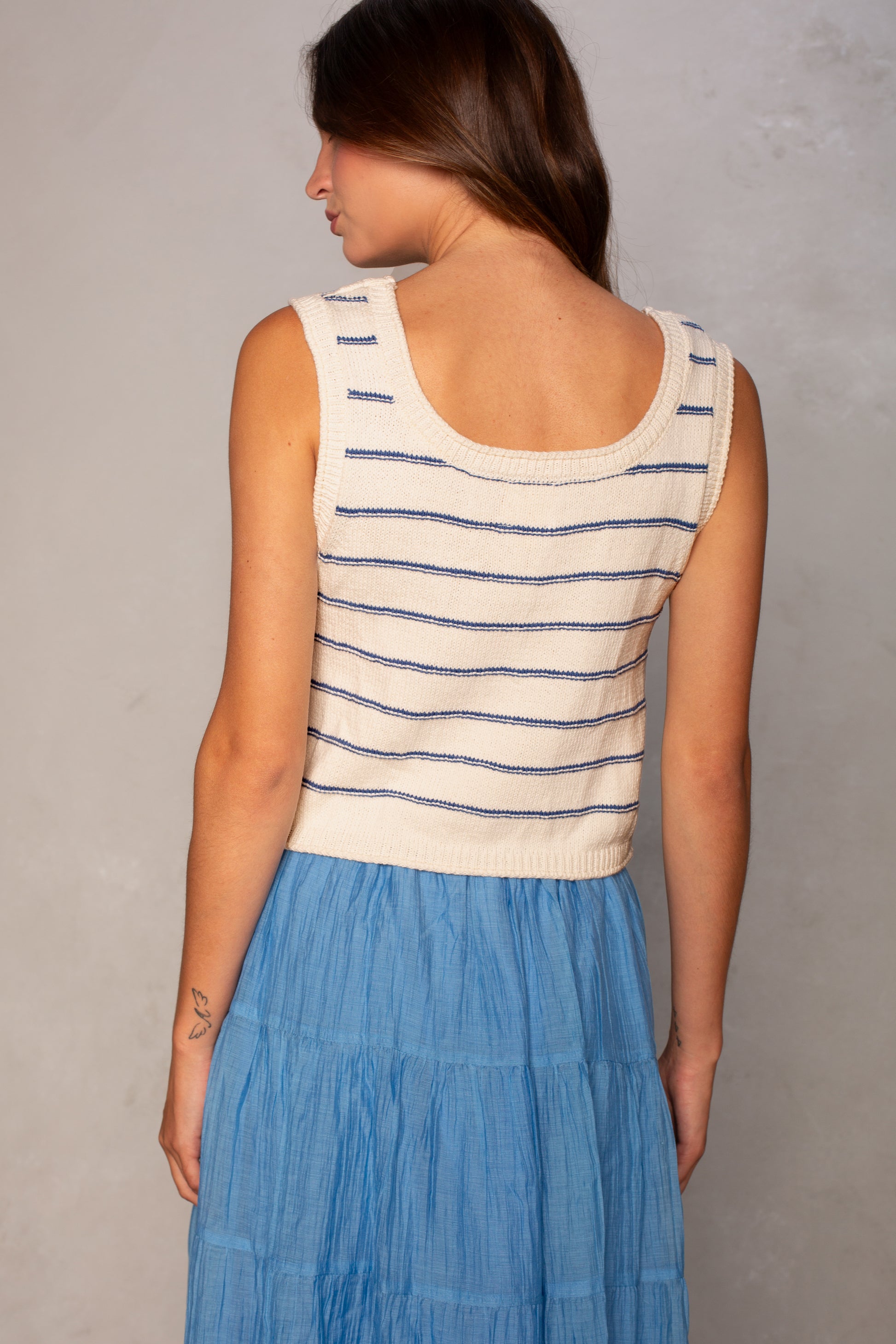 A woman with long brown hair faces away, wearing the Isla Piper Sleeveless Sweater with navy stripes and a light blue tiered skirt. A minimalist tattoo is visible on her right arm against a neutral background.