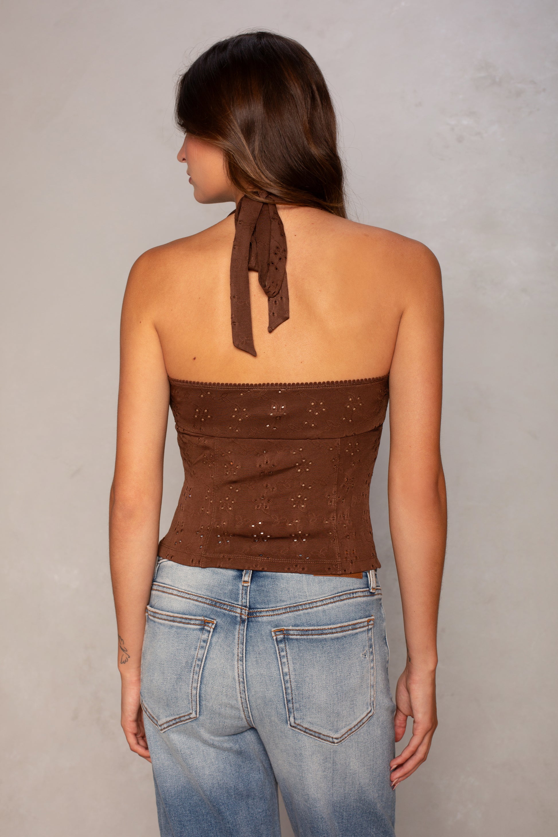 A woman with long brown hair wears the Isla Daisy Eyelet Halter, a cropped brown top, and light blue jeans, standing with her back to the camera against a neutral background.