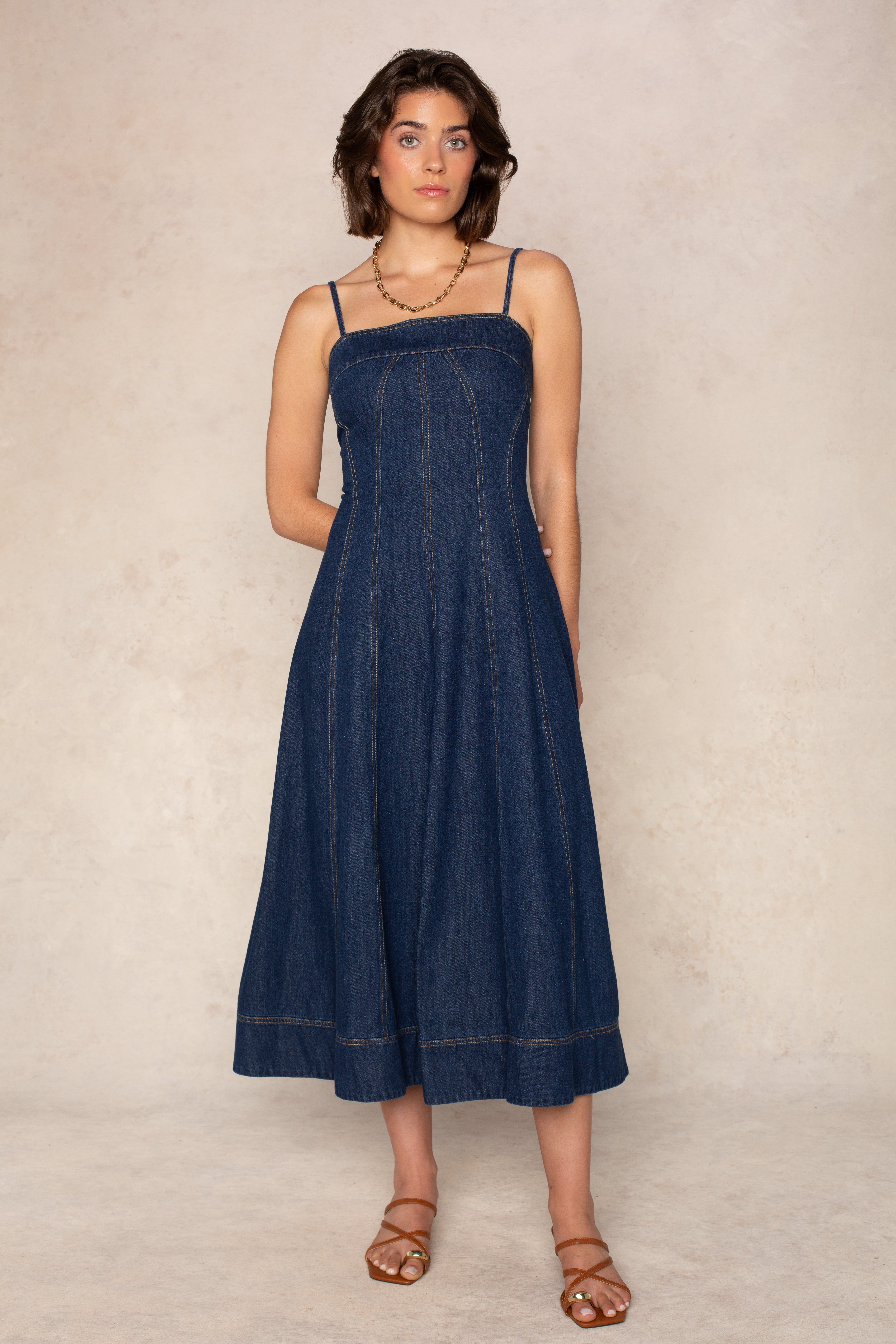 Wearing the Georgie Zoe Denim Midi Dress in Dark Denim, a woman stands against a light beige background. The sleeveless midi dress features a fitted bodice and adjustable spaghetti straps. She pairs the look with brown sandals and a necklace, her hands relaxed by her sides.