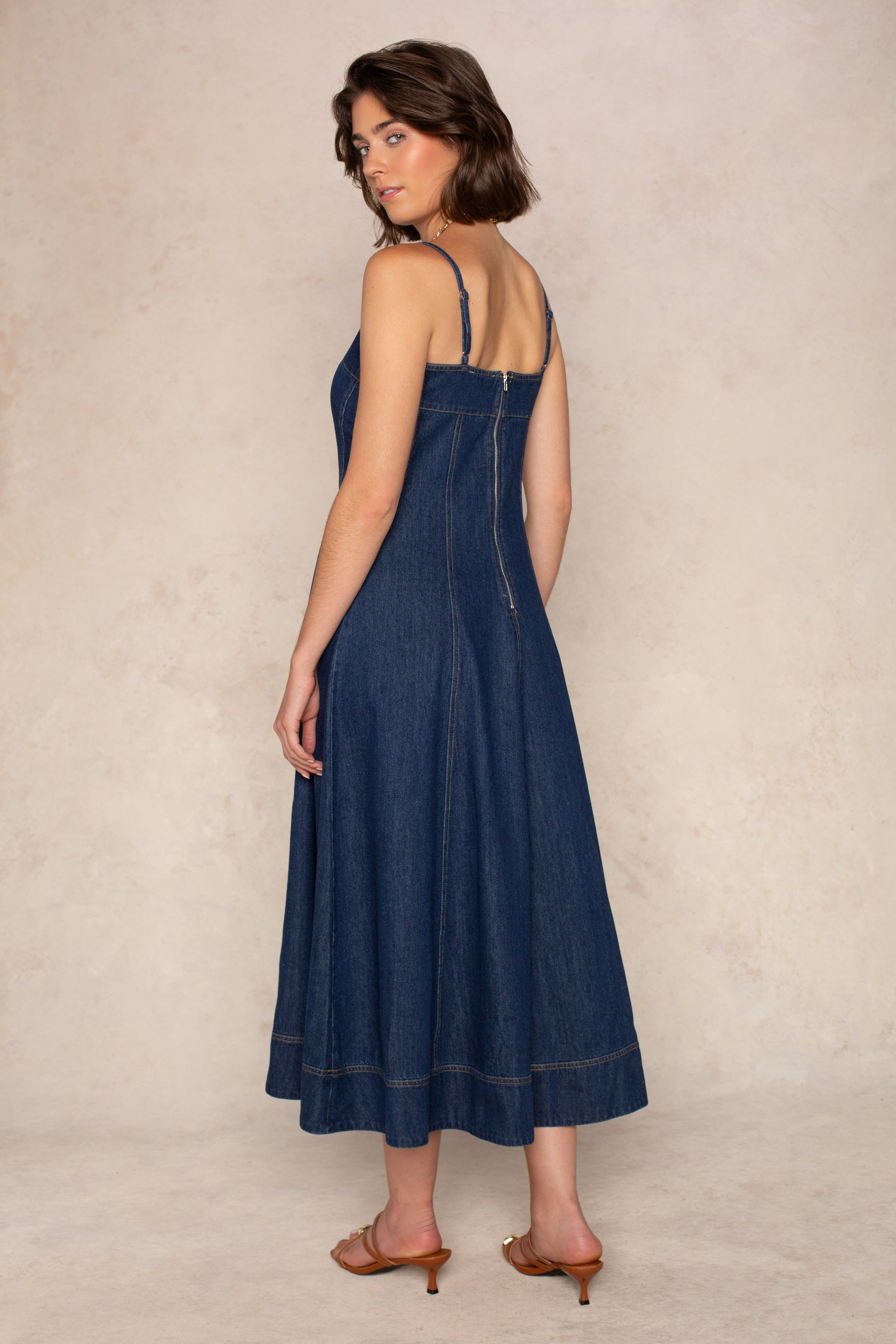 A woman stands on a light background wearing the Georgie Zoe Denim Midi Dress with a fitted bodice and brown heeled sandals, looking over her shoulder at the camera.