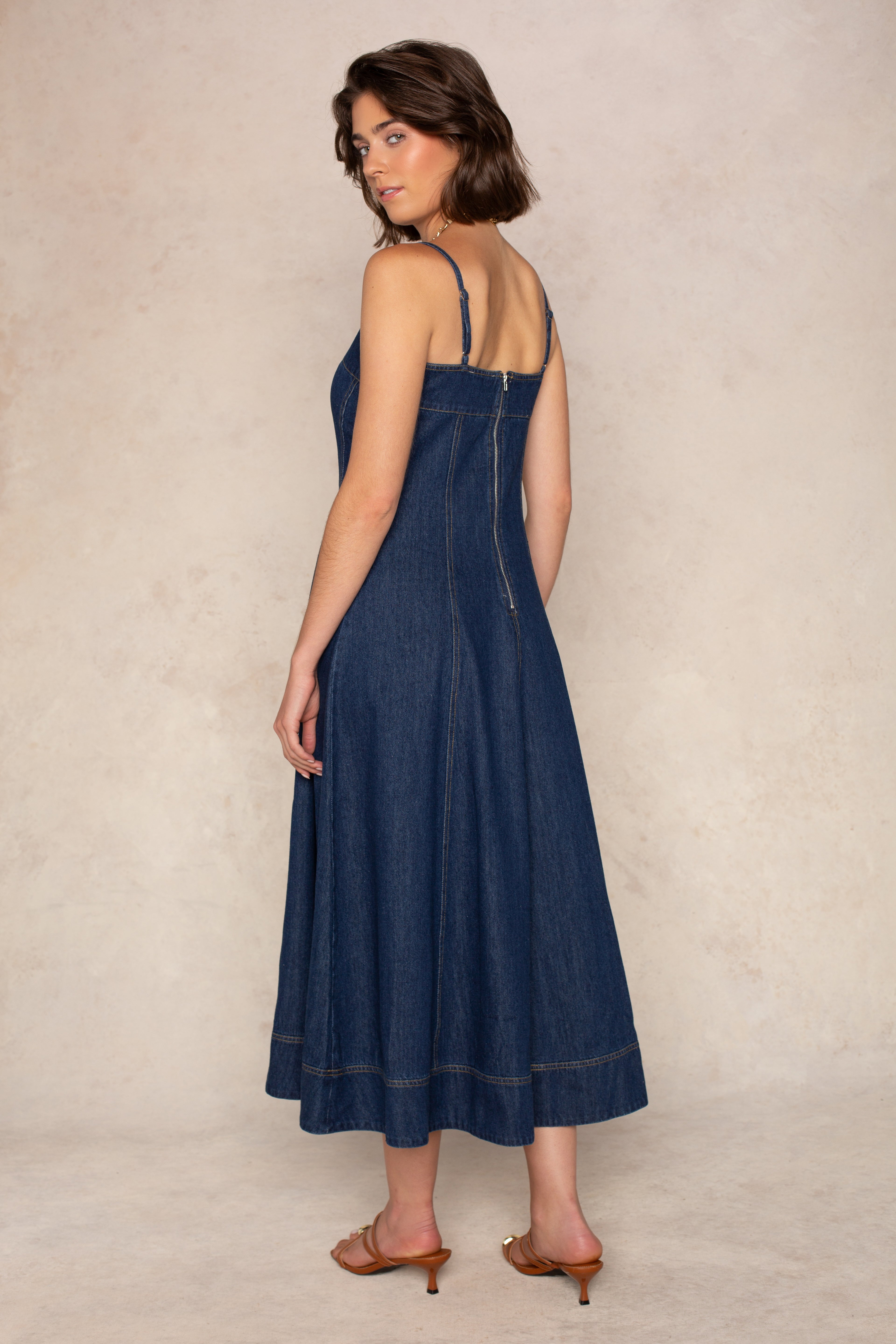 A woman stands on a light background wearing the Georgie Zoe Denim Midi Dress with a fitted bodice and brown heeled sandals, looking over her shoulder at the camera.