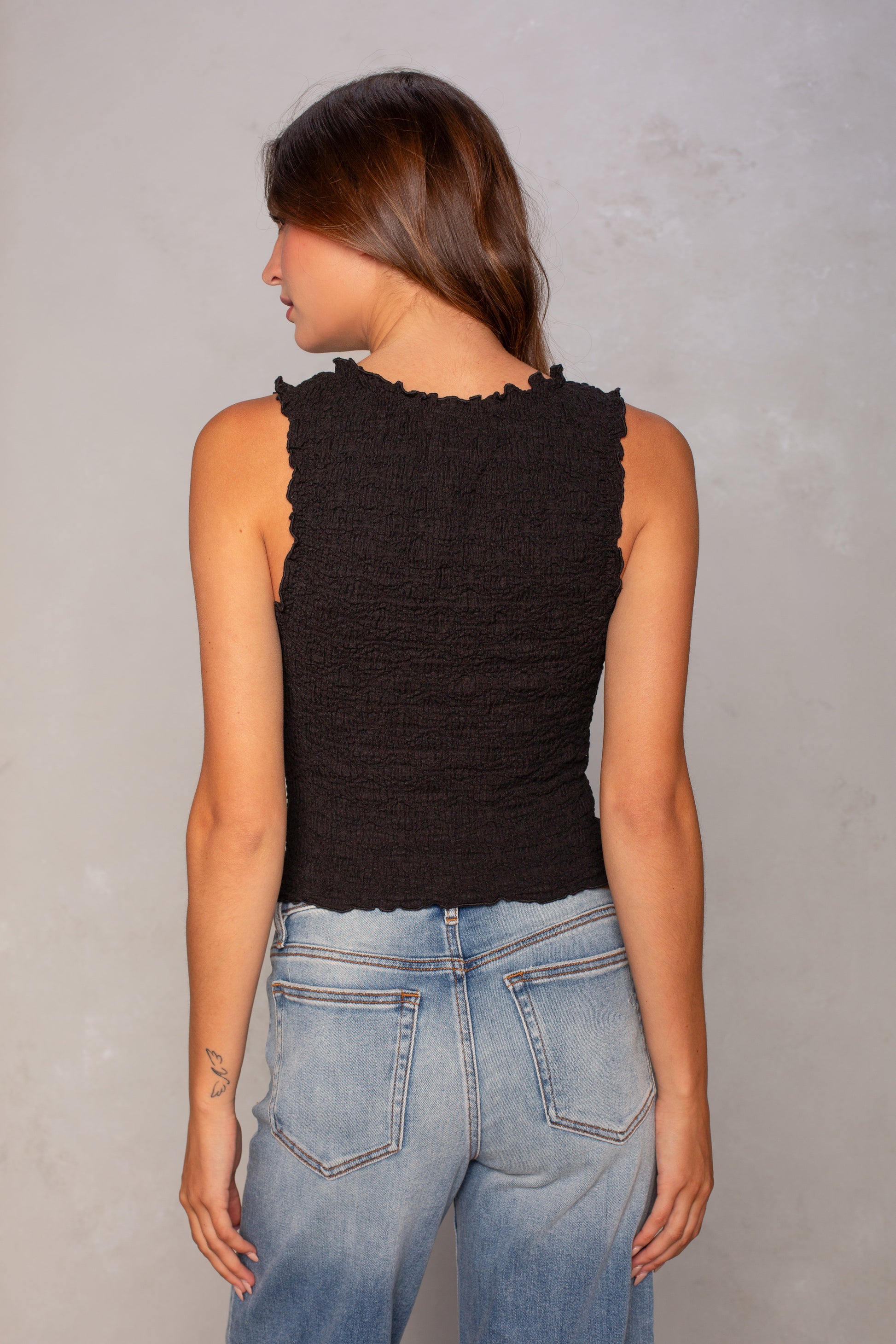 A woman with long brown hair stands facing away against a neutral background, wearing the Isla Ruched Sleeveless Tank in black and light blue jeans.