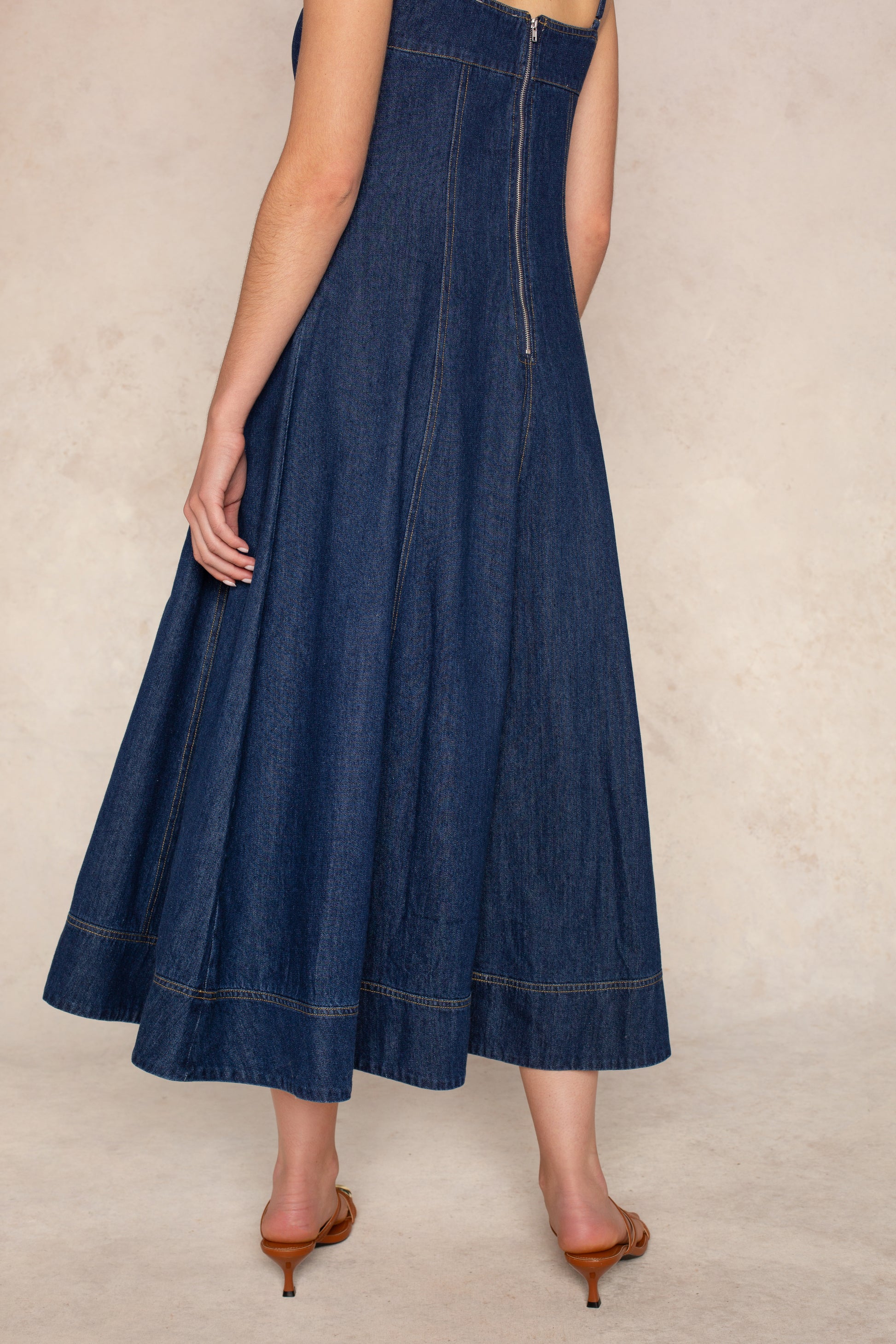 A person wearing the Georgie Zoe Denim Midi Dress—a sleeveless, dark blue denim midi dress with a fitted bodice—paired with brown kitten heels, is shown from the back against a beige background.