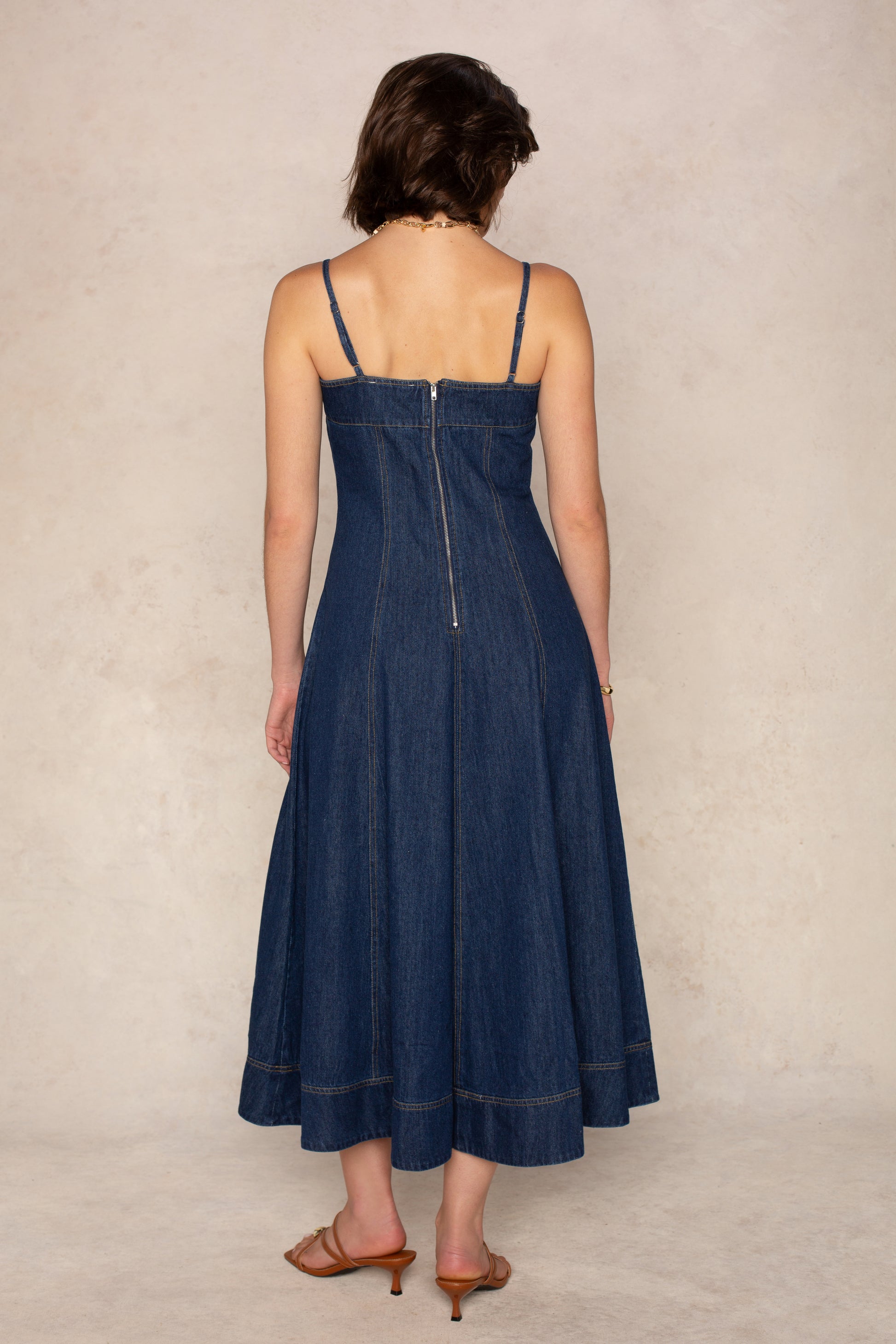 A woman stands with her back to the camera, showcasing the Georgie Zoe Denim Midi Dress—a fitted bodice style with adjustable spaghetti straps and a visible back zipper—paired with brown heeled sandals against a neutral background.
