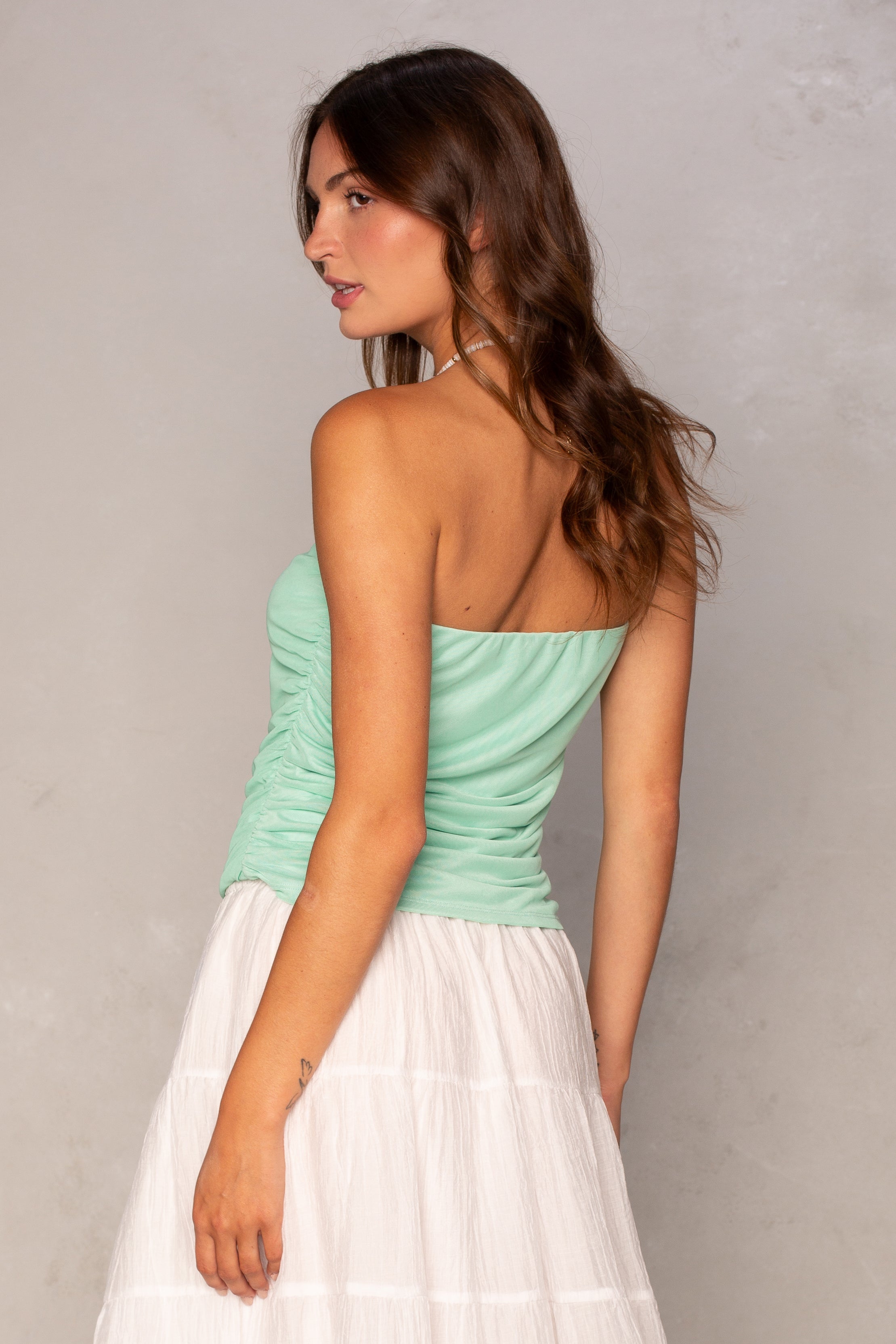 A woman with wavy brown hair models the Isla Mia Mesh Tube, a light green strapless ruched dress with a white tiered skirt. She poses sideways, looking over her shoulder against a neutral light gray background.