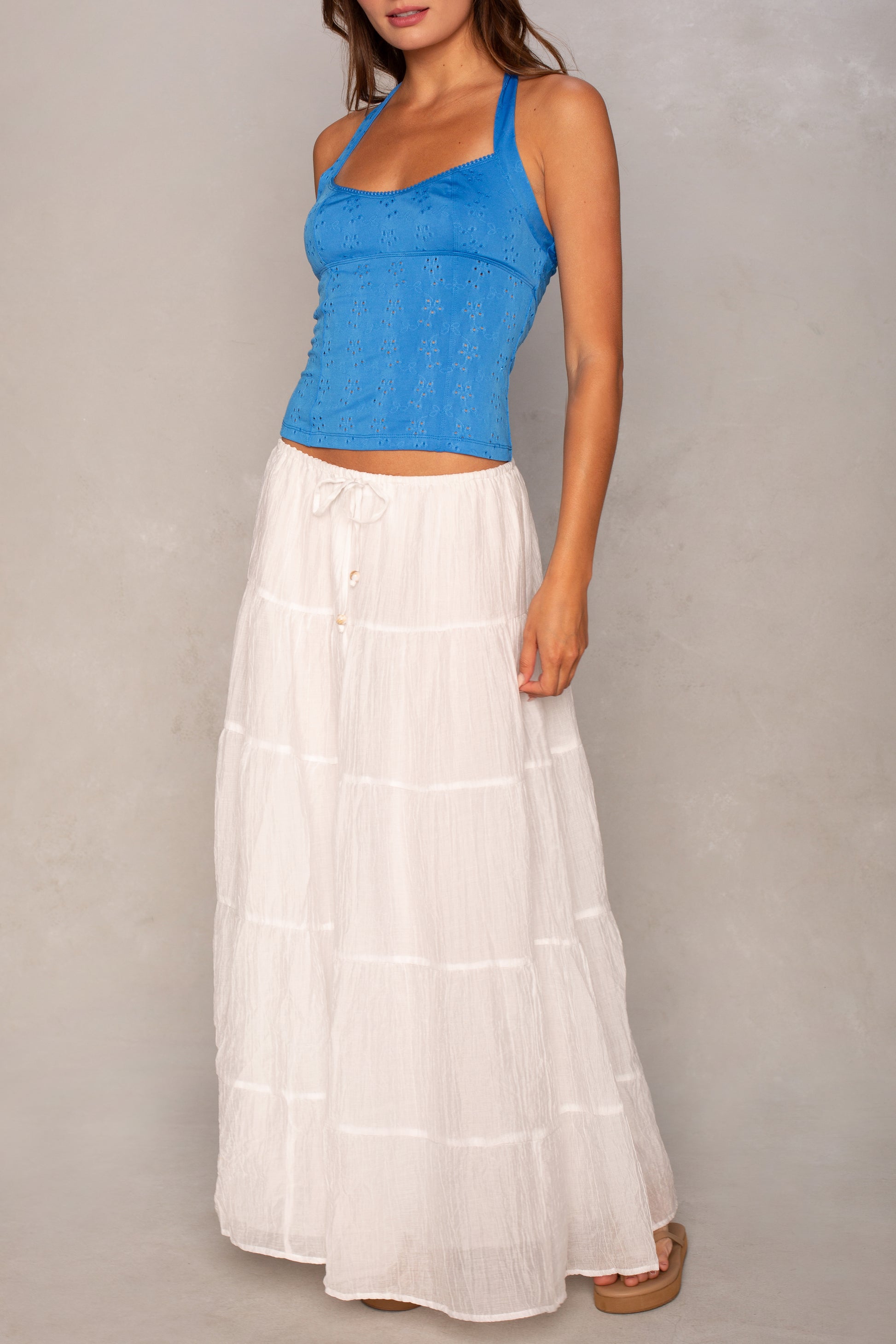 A woman wears the Isla Laney Tiered Maxi Skirt in white with an elastic waistband, paired with a bright blue sleeveless top and sandals, standing against a neutral background.