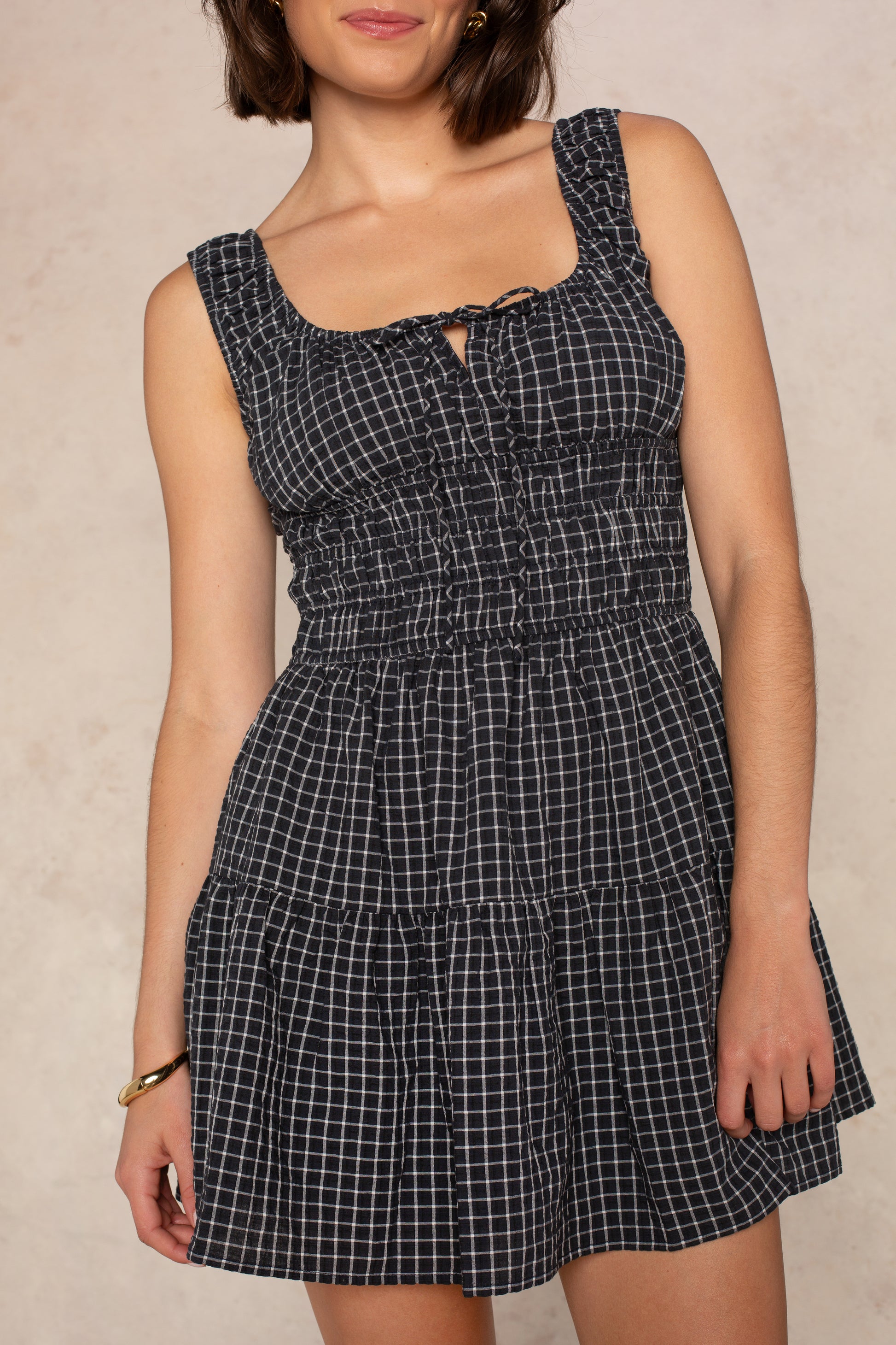 A woman wears the Isla Savannah Mini Dress in Black Multi—a sleeveless dress with a smocked bodice, white checkered pattern, and tiered skirt—standing against a neutral background.