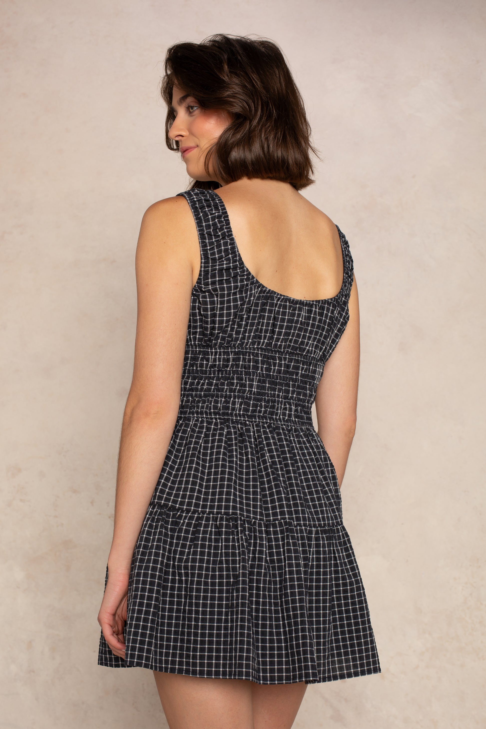 A woman with short brown hair wears the Isla Savannah Mini Dress— a sleeveless black and white checkered dress featuring a scoop neckline and smocked bodice— as she stands with her back to the camera, glancing over her shoulder against a plain beige background.