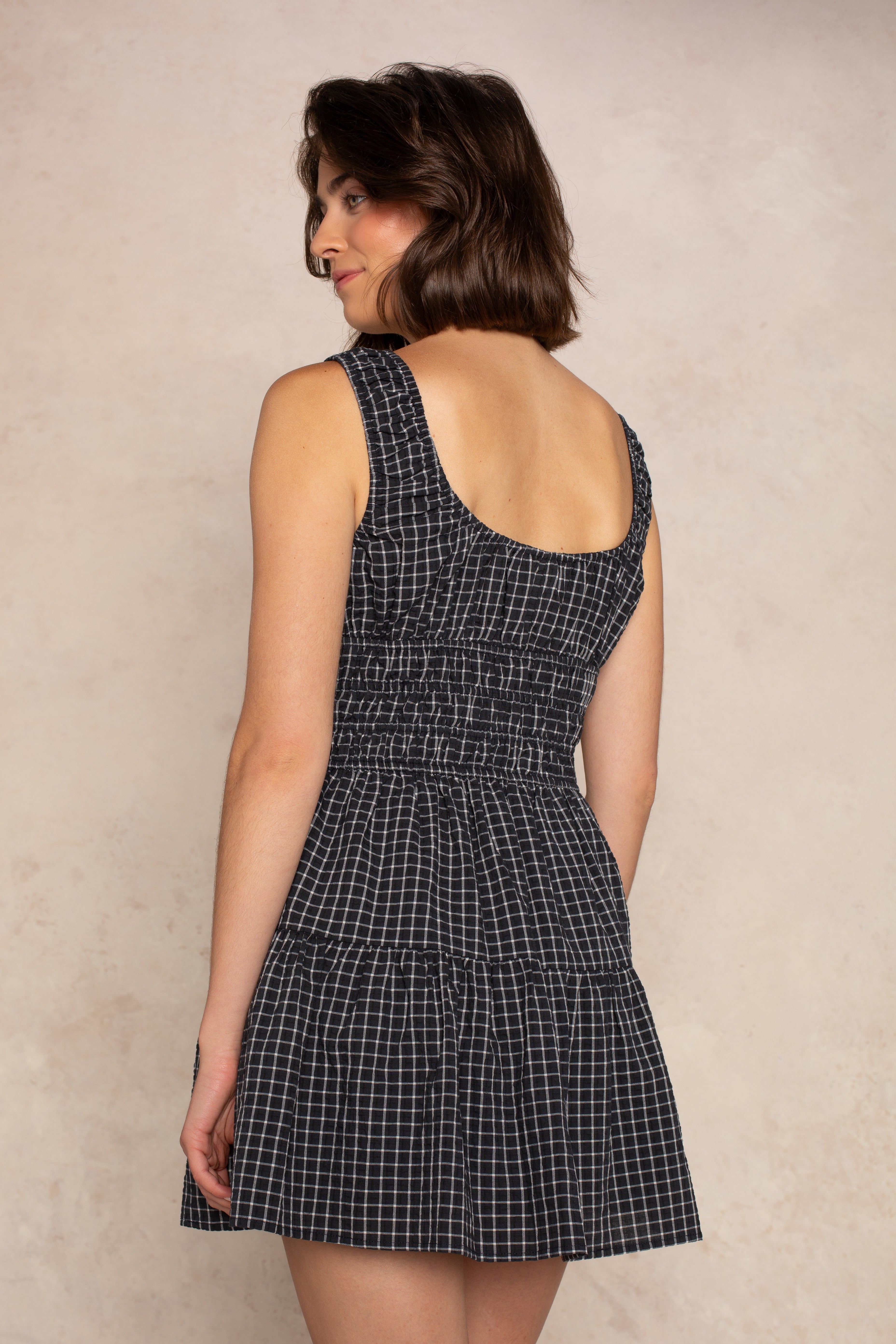 A woman with short brown hair wears the Isla Savannah Mini Dress— a sleeveless black and white checkered dress featuring a scoop neckline and smocked bodice— as she stands with her back to the camera, glancing over her shoulder against a plain beige background.