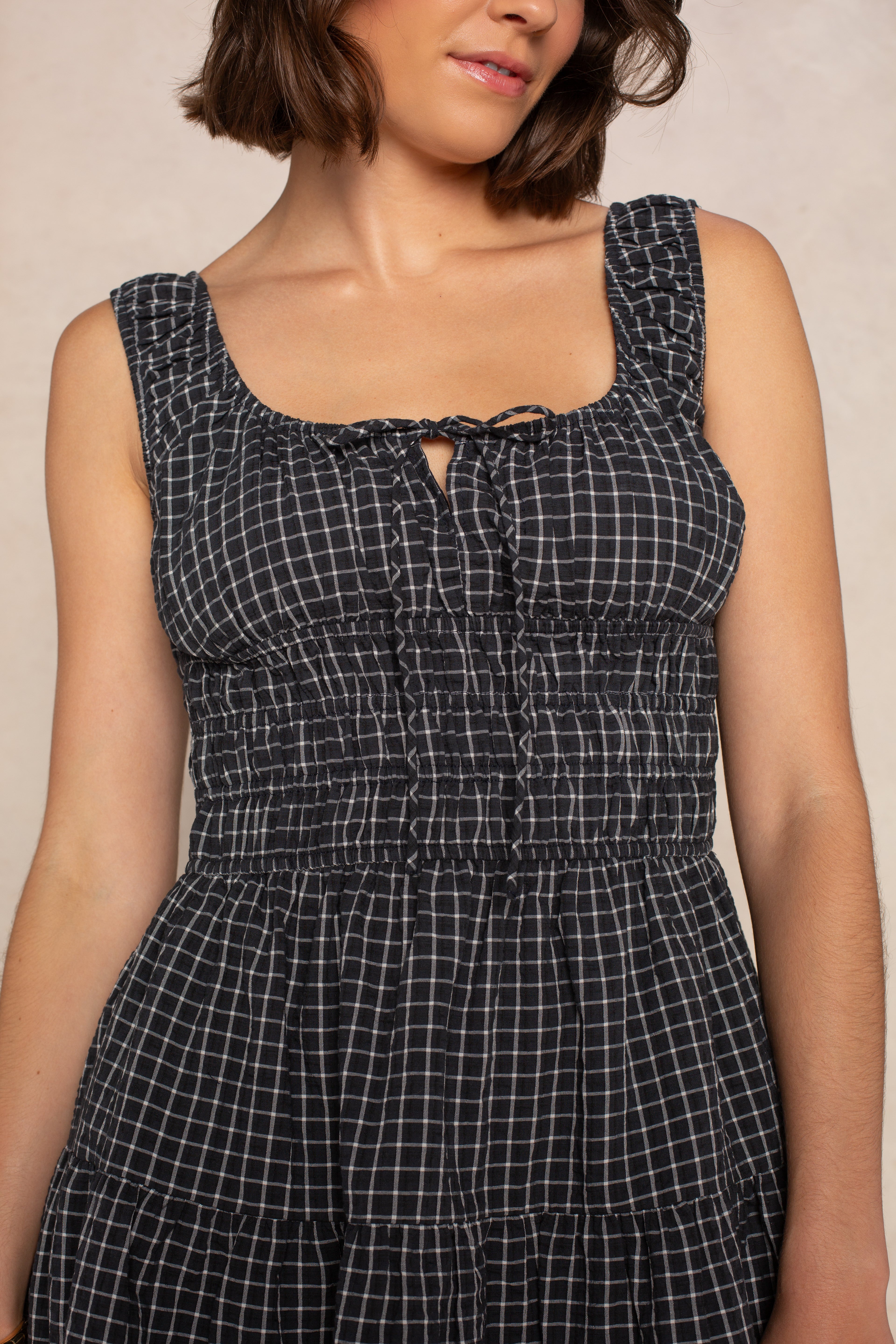 A woman wearing the Isla Savannah Mini Dress—a sleeveless black mini dress with a white checkered pattern, tie neckline, and smocked bodice—shown from shoulders to waist.