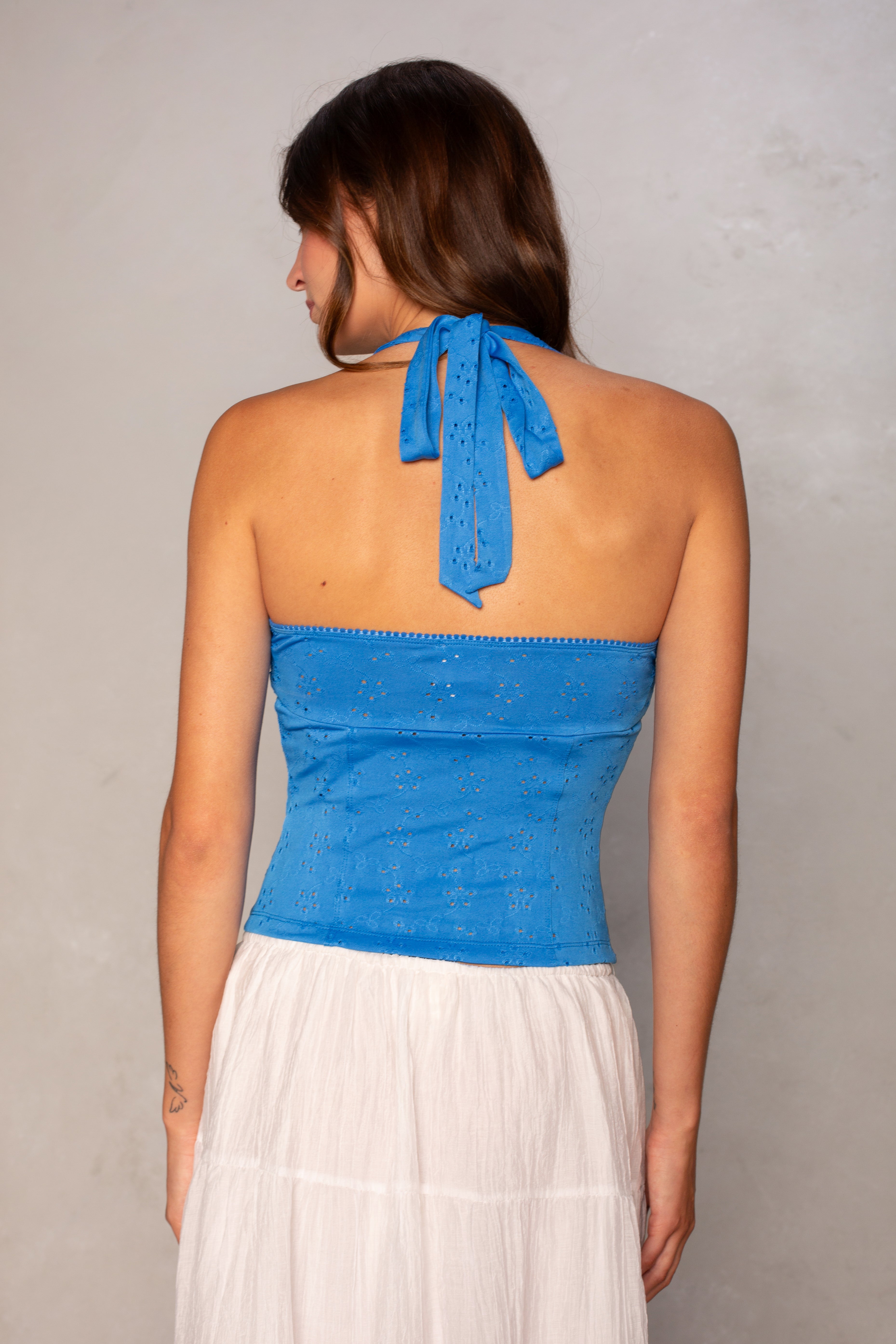 A woman with long brown hair is shown from the back, wearing the Isla Daisy Eyelet Halter in blue and a white skirt. The cropped top stands out with its modern style against a plain, light-colored background.