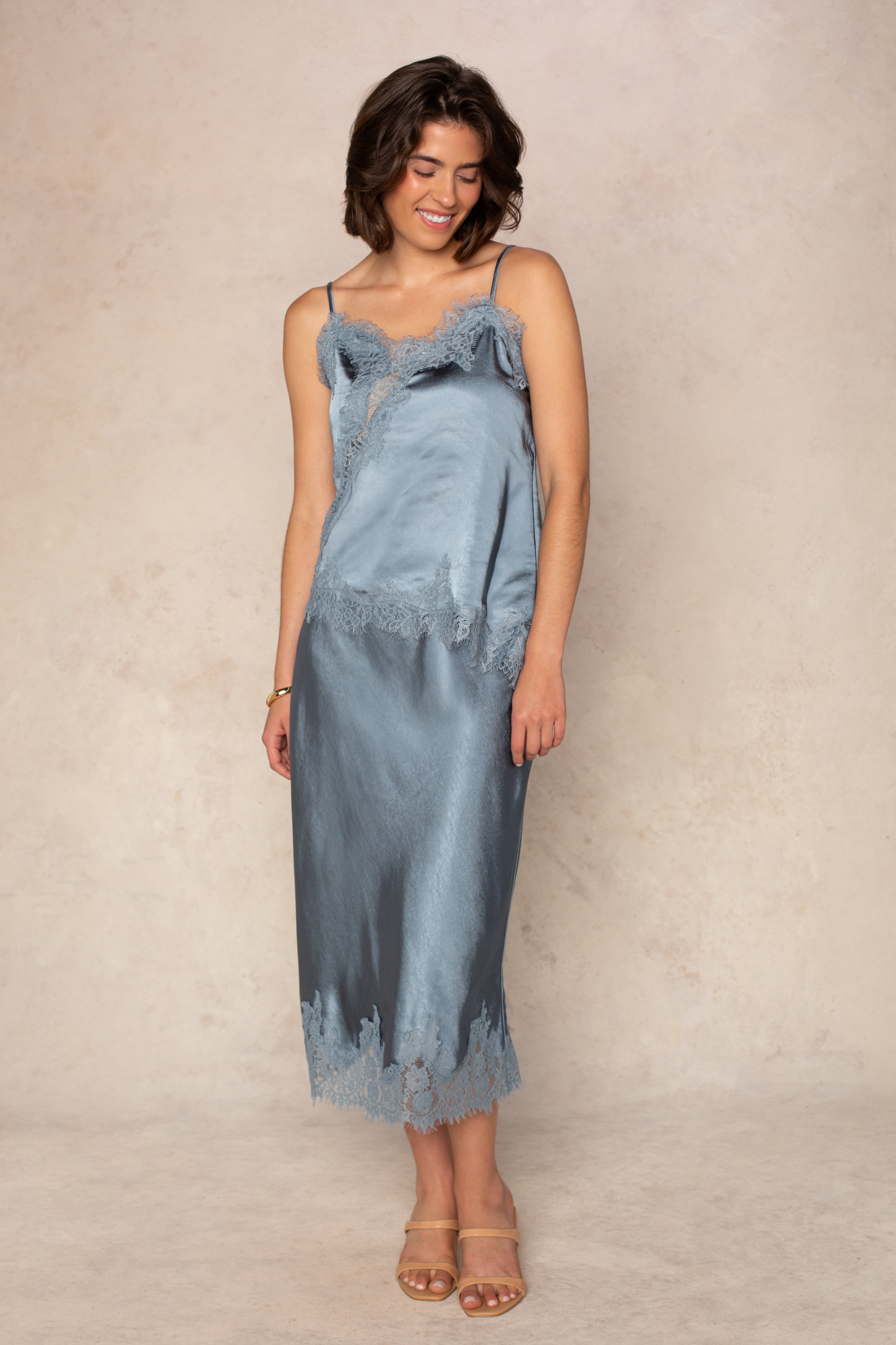 A woman stands against a neutral background, smiling with her head slightly tilted and her hands relaxed at her sides. She is wearing the Georgie Remi Asym Lace Satin Cami, a silky, light blue cami featuring lace trim and adjustable spaghetti straps, paired with strappy nude heels.