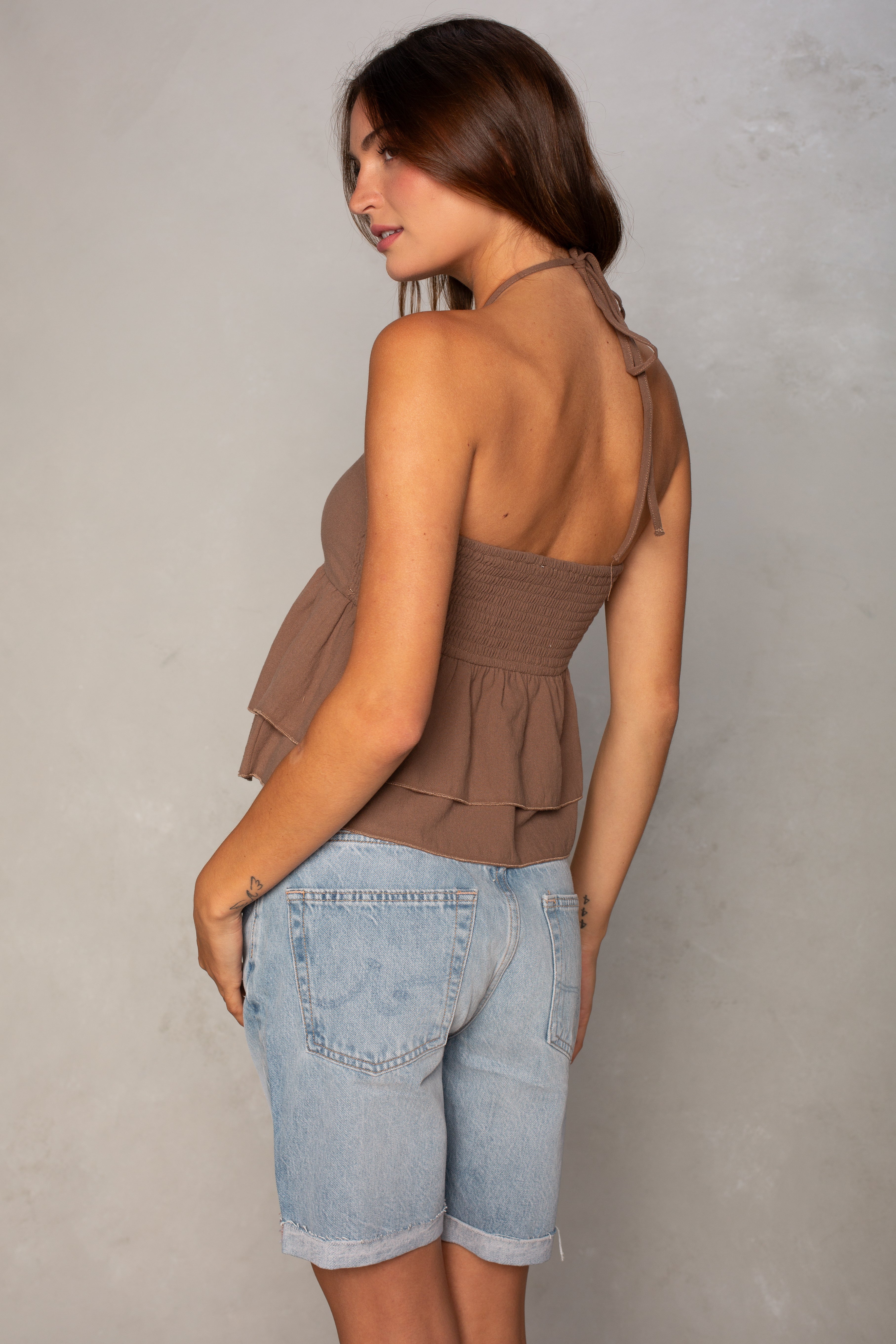 A woman with long brown hair models the Isla Josie Textured Halter Top in brown, paired with light blue denim shorts, standing sideways and looking over her shoulder against a light gray background.