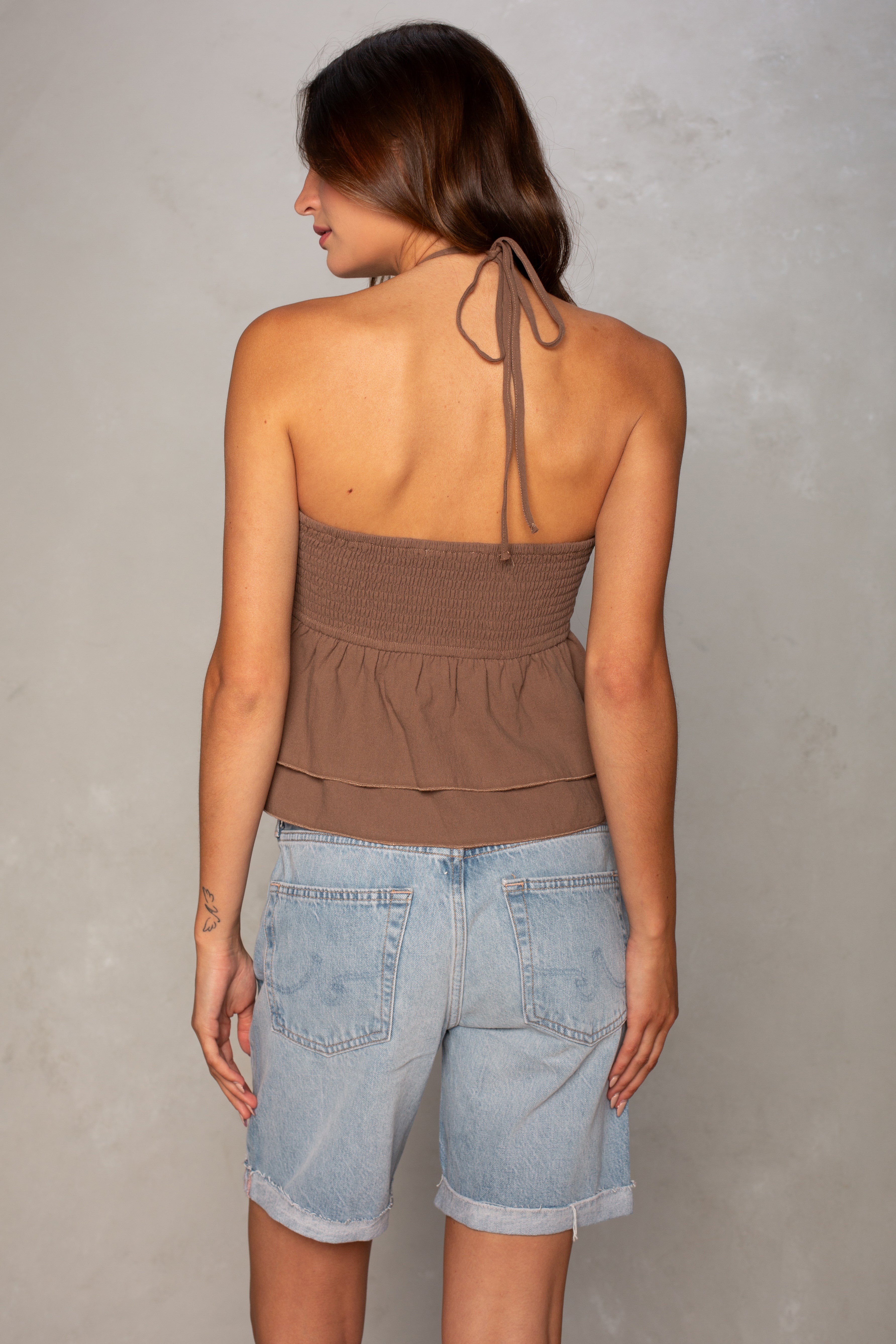 A woman with long brown hair models the Isla Josie Textured Halter Top in brown, paired with light blue denim shorts, standing with her back to the camera against a plain light gray background.