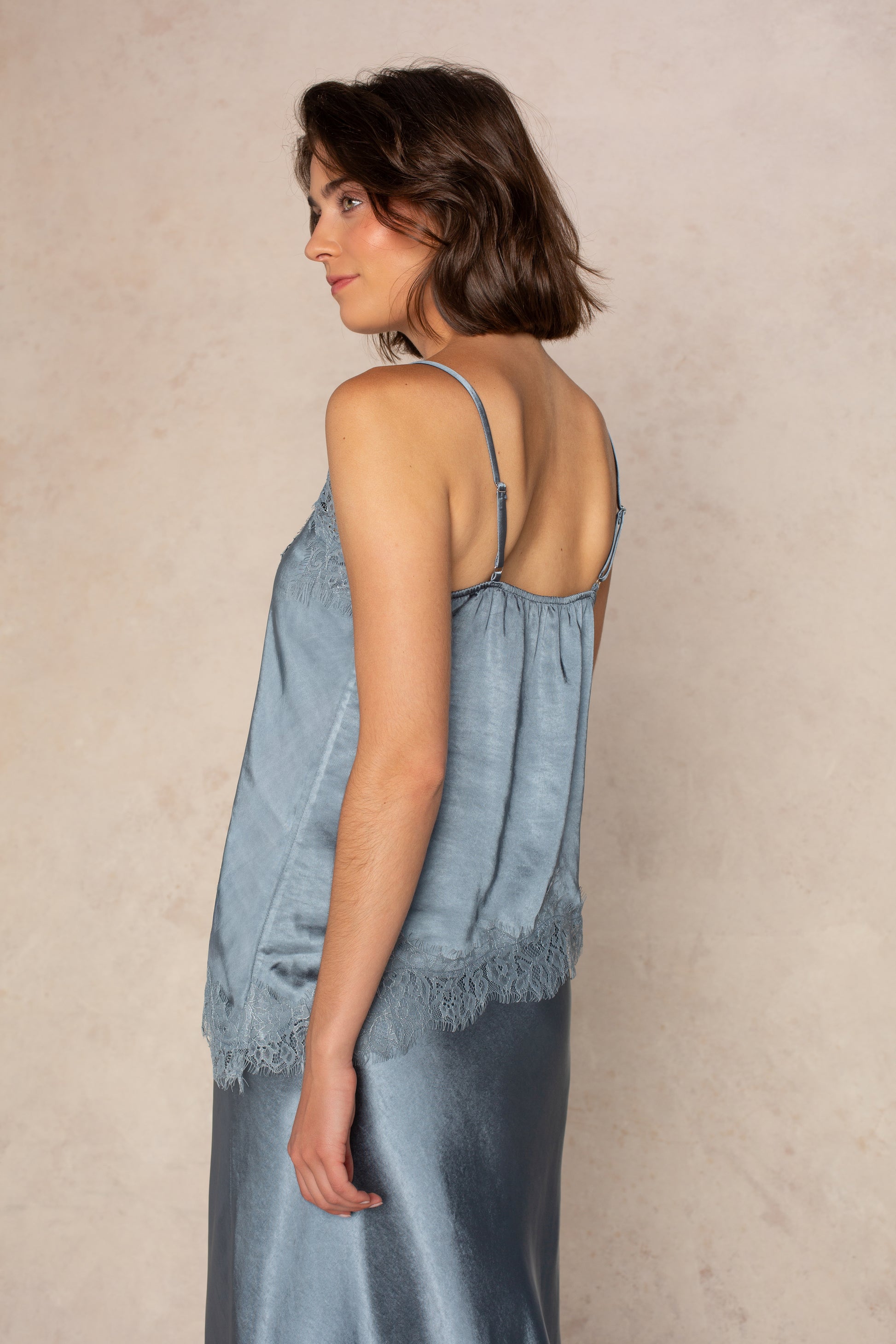 A woman wearing the Georgie Remi Asym Lace Satin Cami—a light blue satin top with delicate lace trim—paired with a matching skirt, stands sideways against a beige background, looking over her shoulder.