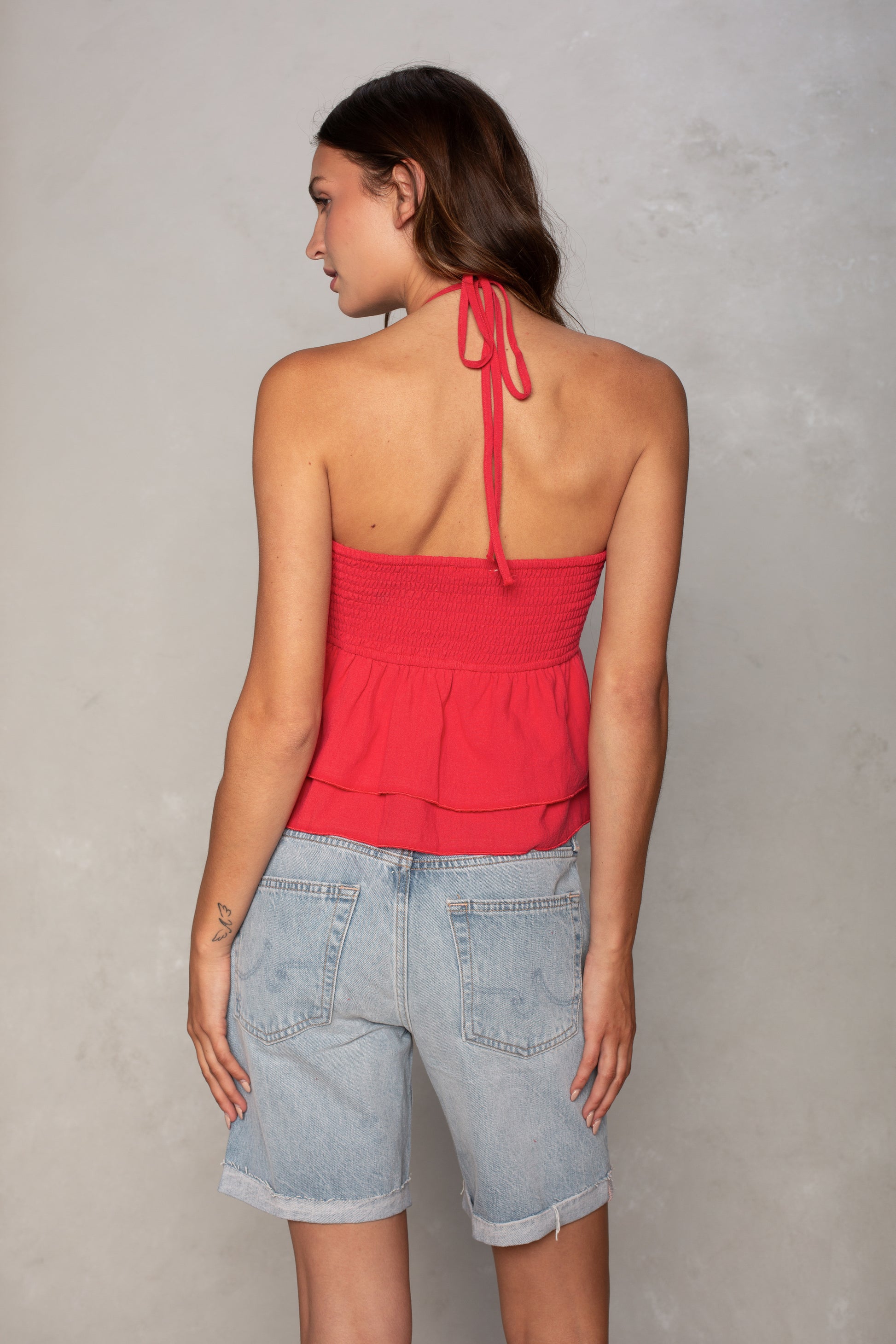 A woman with straight brown hair faces away from the camera, wearing the Isla Josie Textured Halter Top in red and light blue denim shorts, her left hand resting on her thigh against a plain light gray wall.