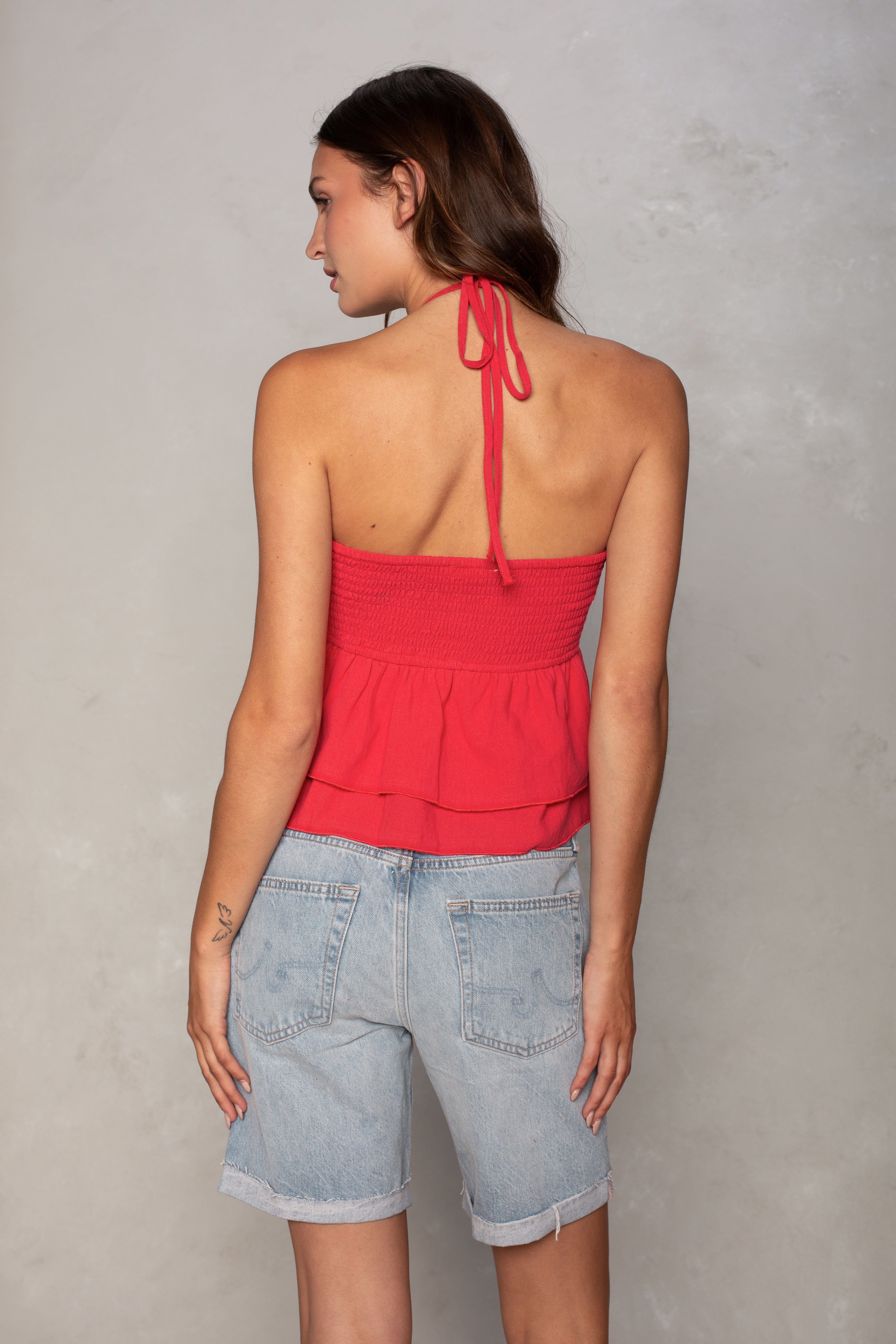 A woman with straight brown hair faces away from the camera, wearing the Isla Josie Textured Halter Top in red and light blue denim shorts, her left hand resting on her thigh against a plain light gray wall.