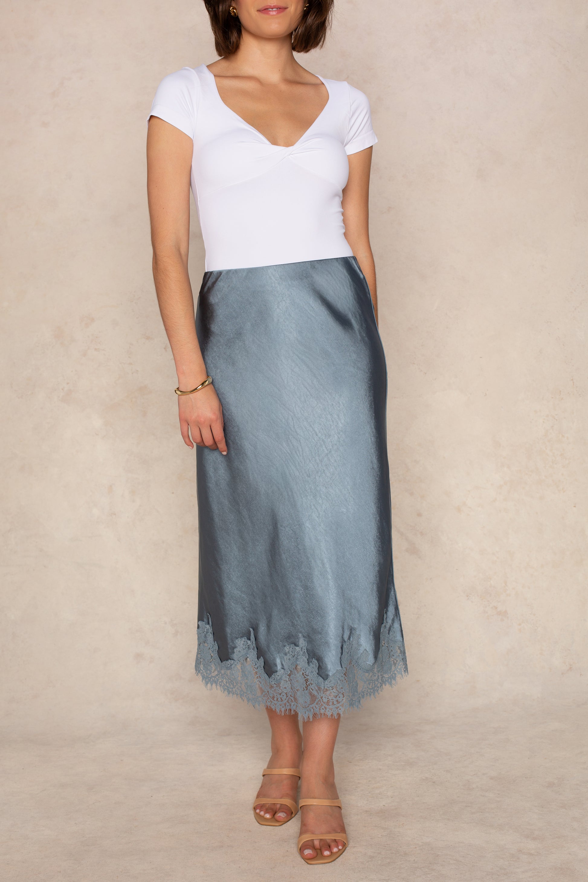 A woman models the Georgie Remi Midi Lace Satin Skirt in SLATE BLUE, featuring a chic bias-cut silhouette and delicate lace trim at the hem. She pairs it with a white short-sleeve top, tan sandals, and a gold bracelet while standing against a beige background.