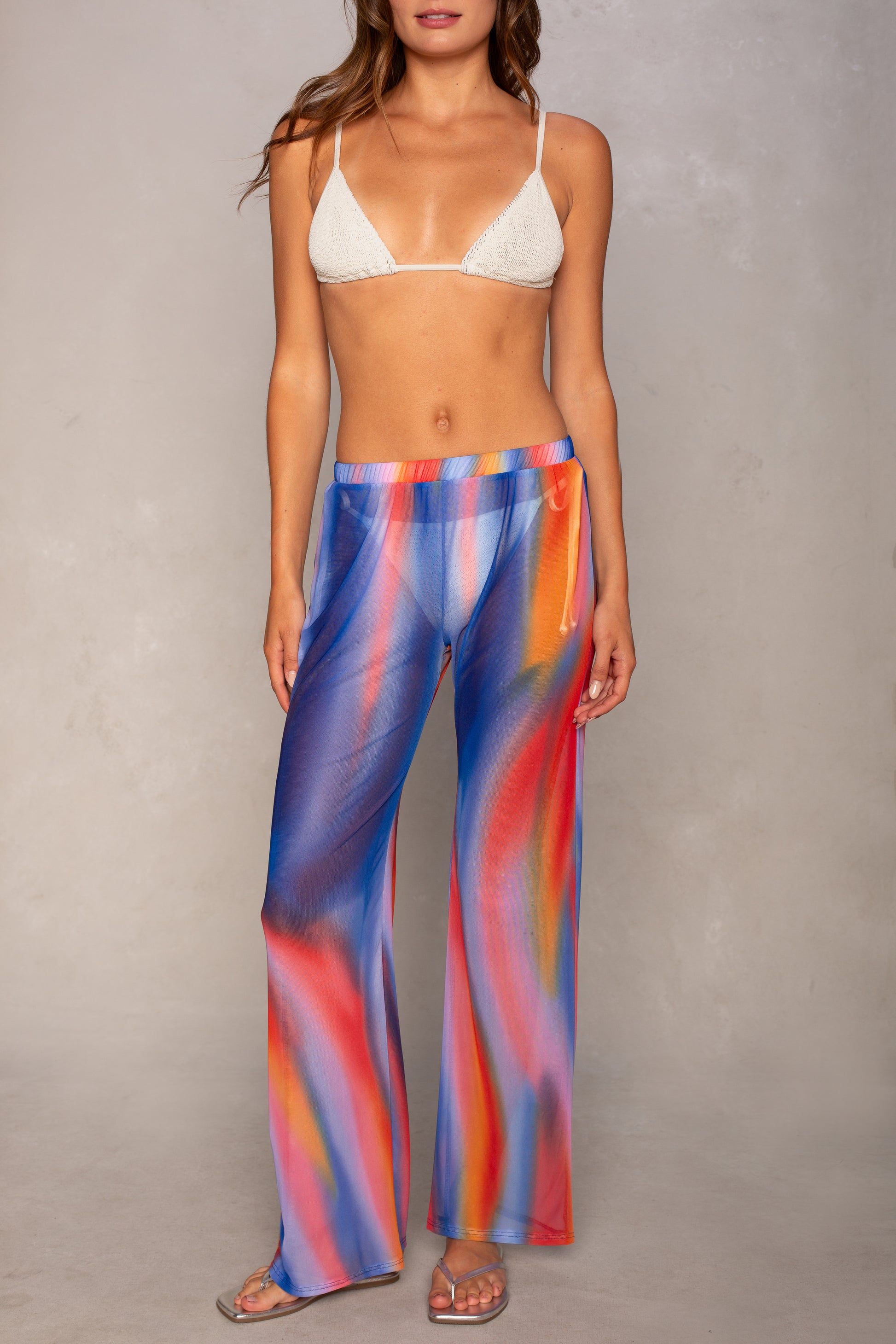 An Isla Mandy Mesh Pant Coverup, featuring a multicolor abstract print in shades of blue, red, yellow, and orange, is styled here with a white bikini top for the perfect beach ensemble against a gray wall.