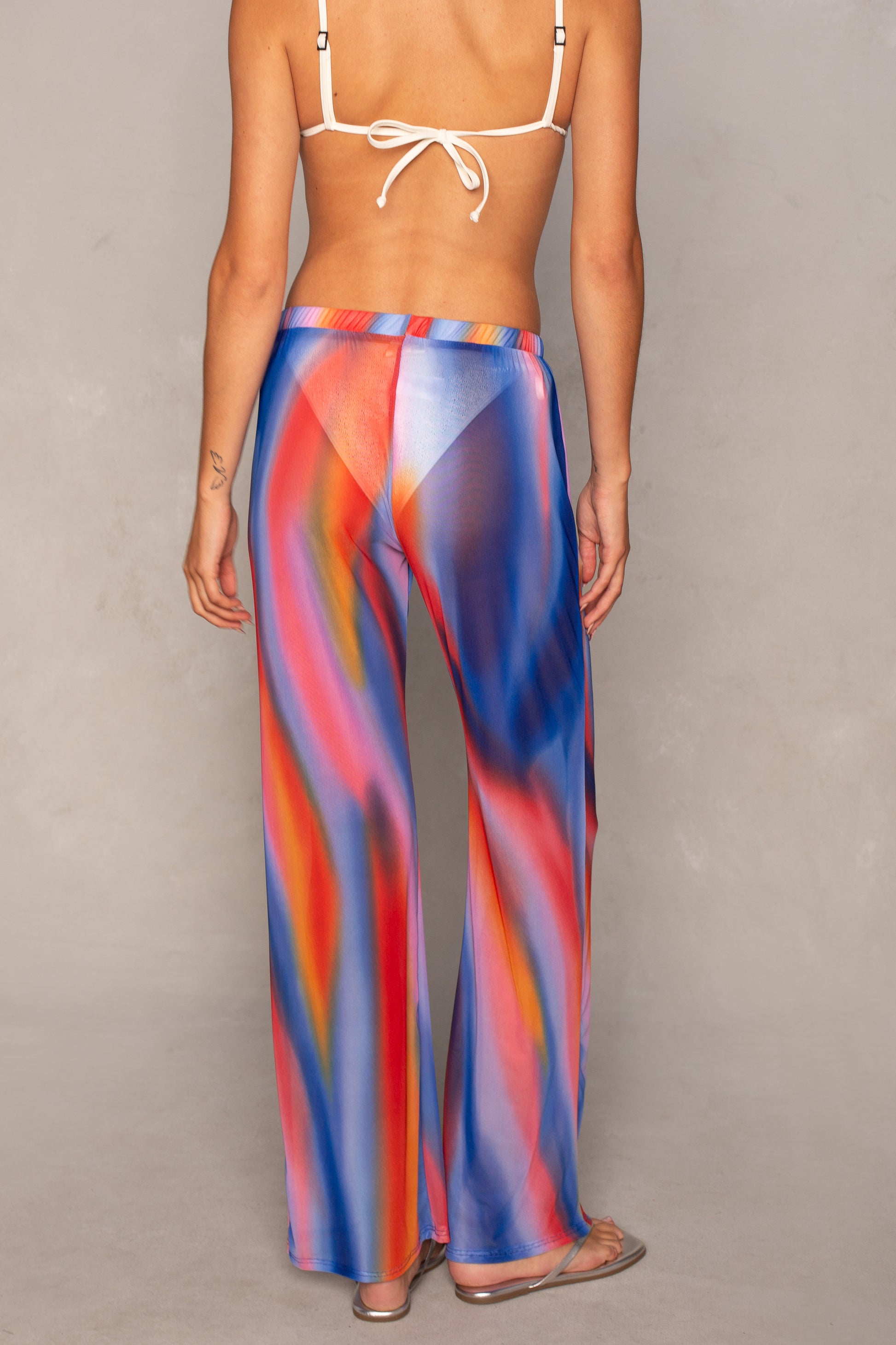 A person stands with their back to the camera, barefoot on a light background, wearing Isla’s Mandy Mesh Pant Coverup—a colorful, sheer, wide-leg pant with a swirling rainbow pattern—and a white bikini top.