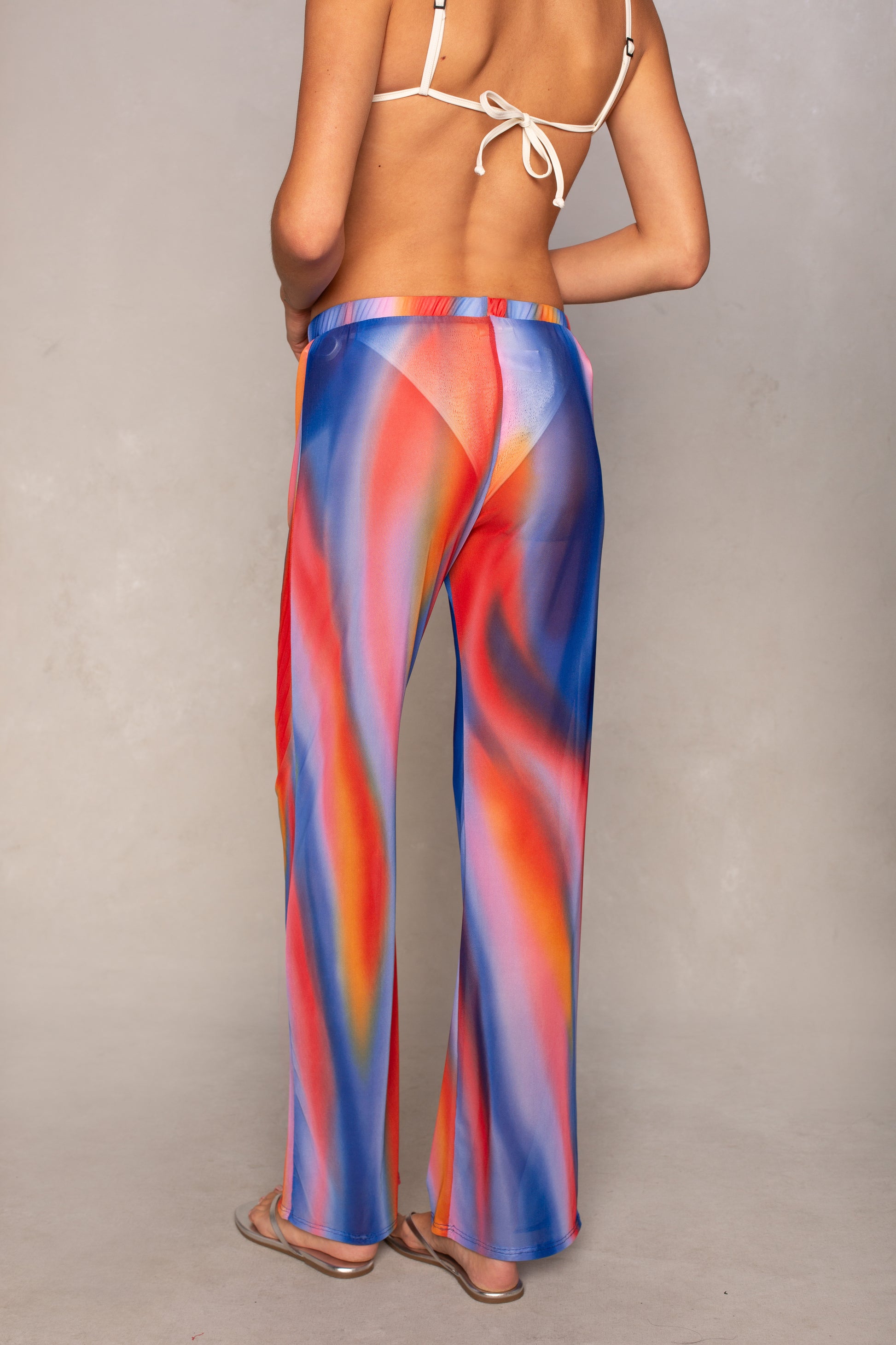 A person models the Isla Mandy Mesh Pant Coverup—colorful, flowy, high-waisted pants with a vibrant swirling pattern—paired with a white bikini top and flip-flops, standing against a plain background.