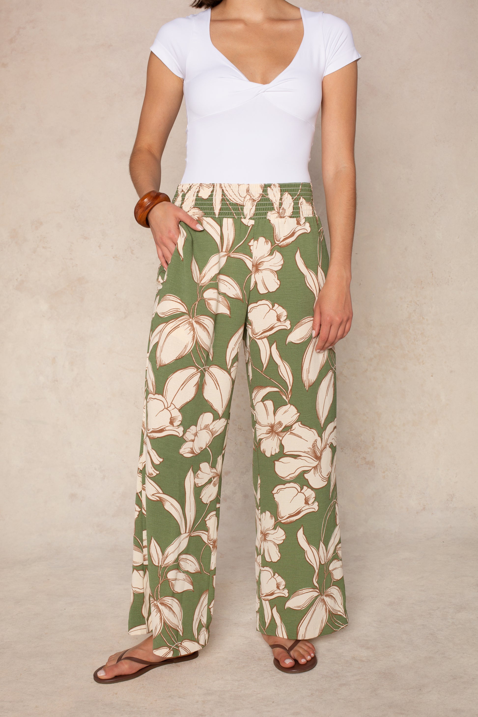 A woman models the Isla Hattie Beach Pant in green—a tropical floral wide-leg style with a smocked waistband—paired with a white short-sleeve top, brown bangle, and sandals. Her face is not shown; background is neutral.