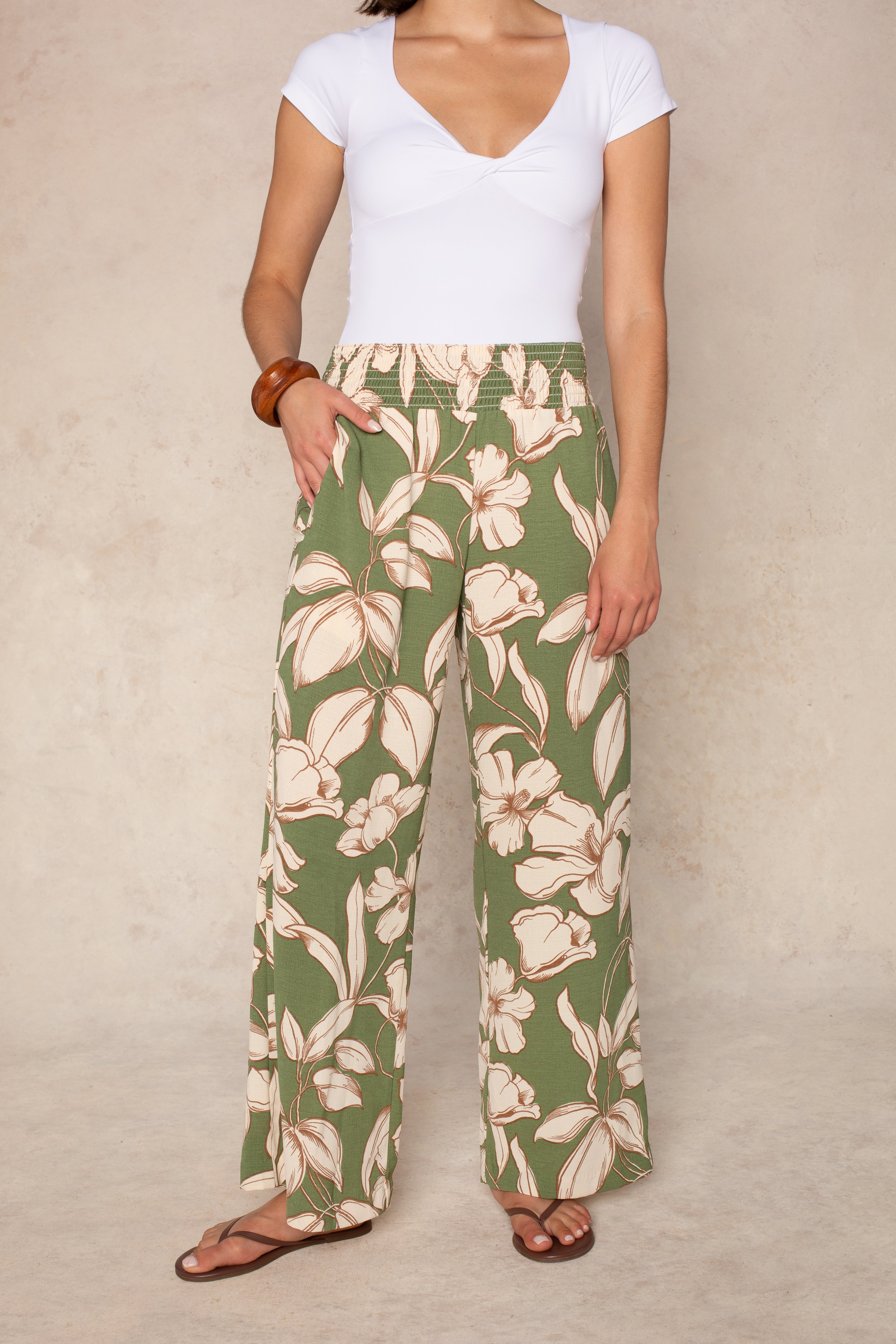 A woman models the Isla Hattie Beach Pant in green—a tropical floral wide-leg style with a smocked waistband—paired with a white short-sleeve top, brown bangle, and sandals. Her face is not shown; background is neutral.