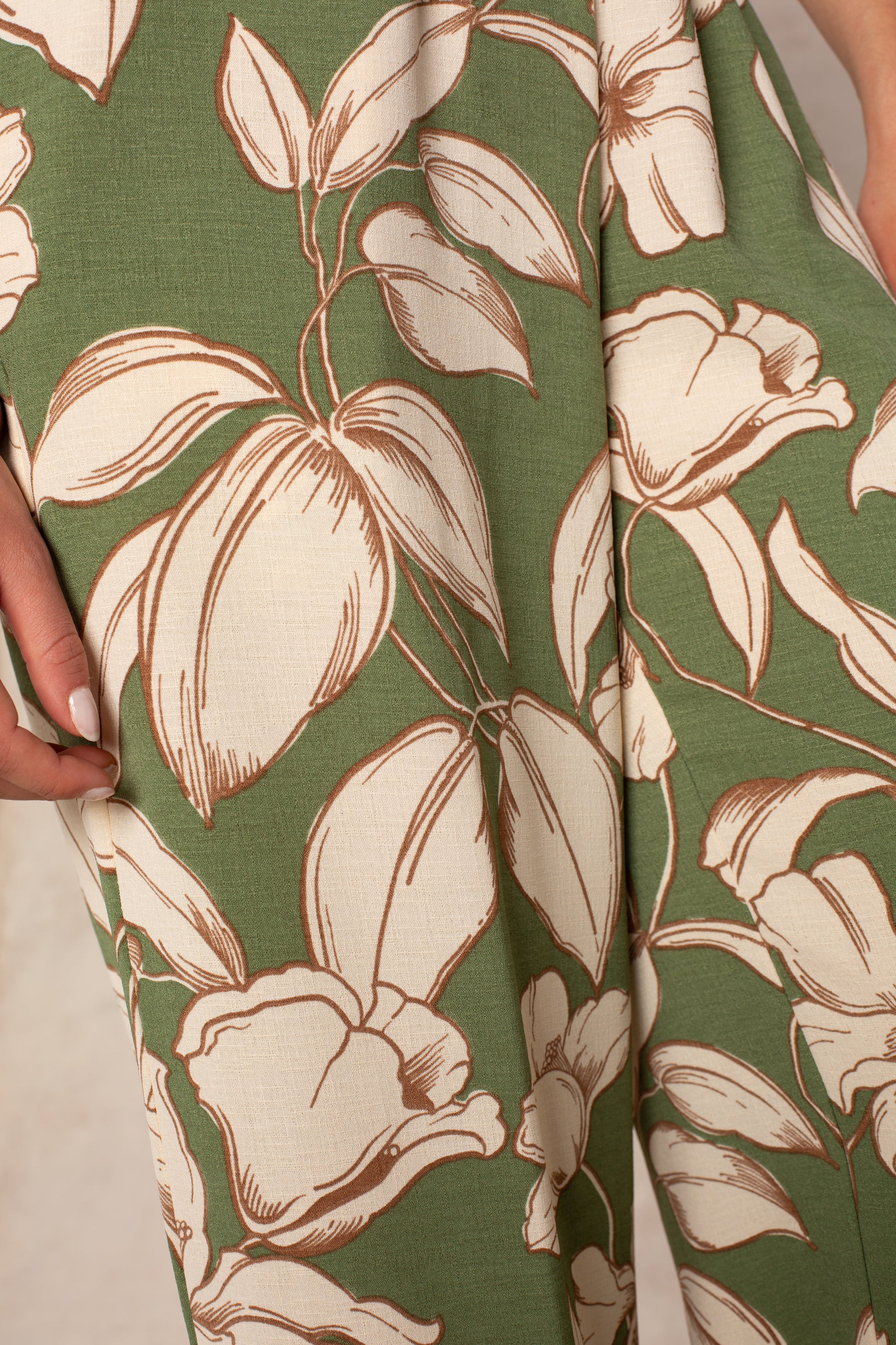 Close-up of Isla's Hattie Beach Pant: green fabric with bold cream and brown tropical florals. A manicured hand touches the material, evoking the comfort and style of its wide-leg cut and smocked waistband.