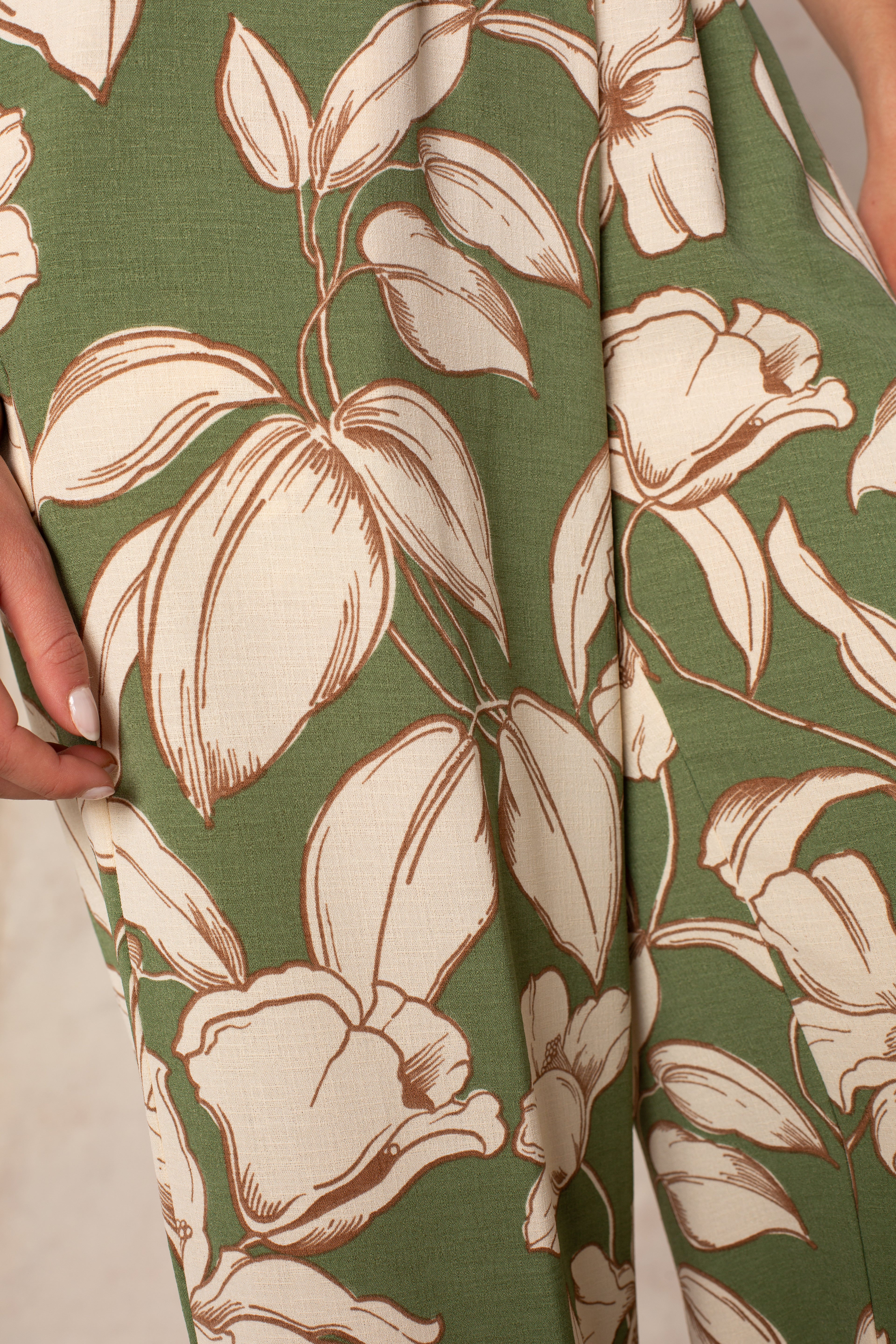 Close-up of Isla's Hattie Beach Pant: green fabric with bold cream and brown tropical florals. A manicured hand touches the material, evoking the comfort and style of its wide-leg cut and smocked waistband.