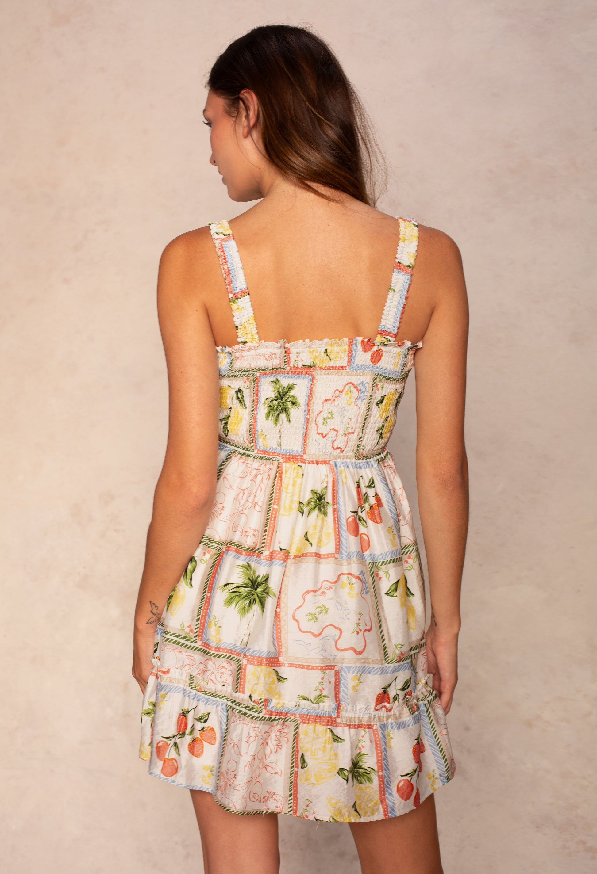 A woman is seen from behind in the Georgie Cece Smocked Mini Dress, featuring a smocked bodice, ruffled straps, and a tiered mini skirt with colorful tropical fruit and palm tree prints on a neutral beige background.