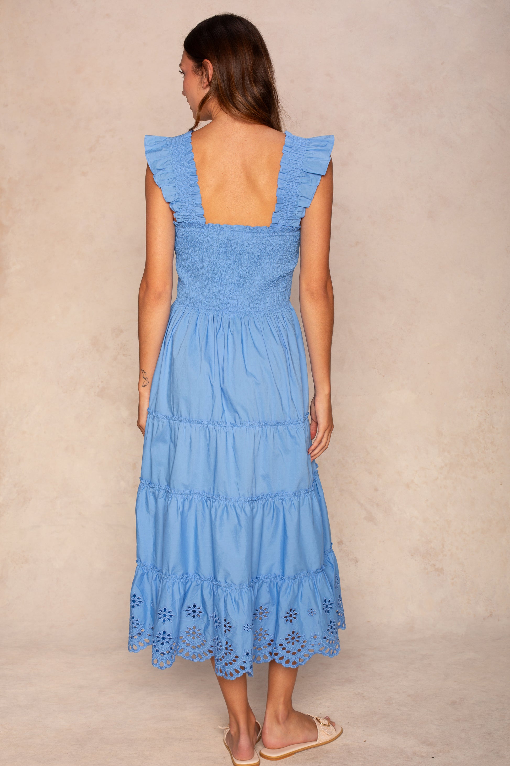 A woman faces away, wearing the Georgie Calypso Smocked Scallop Midi Dress in light blue with ruffled straps, a smocked bodice, scalloped eyelet hem, and flat sandals against a light neutral background.