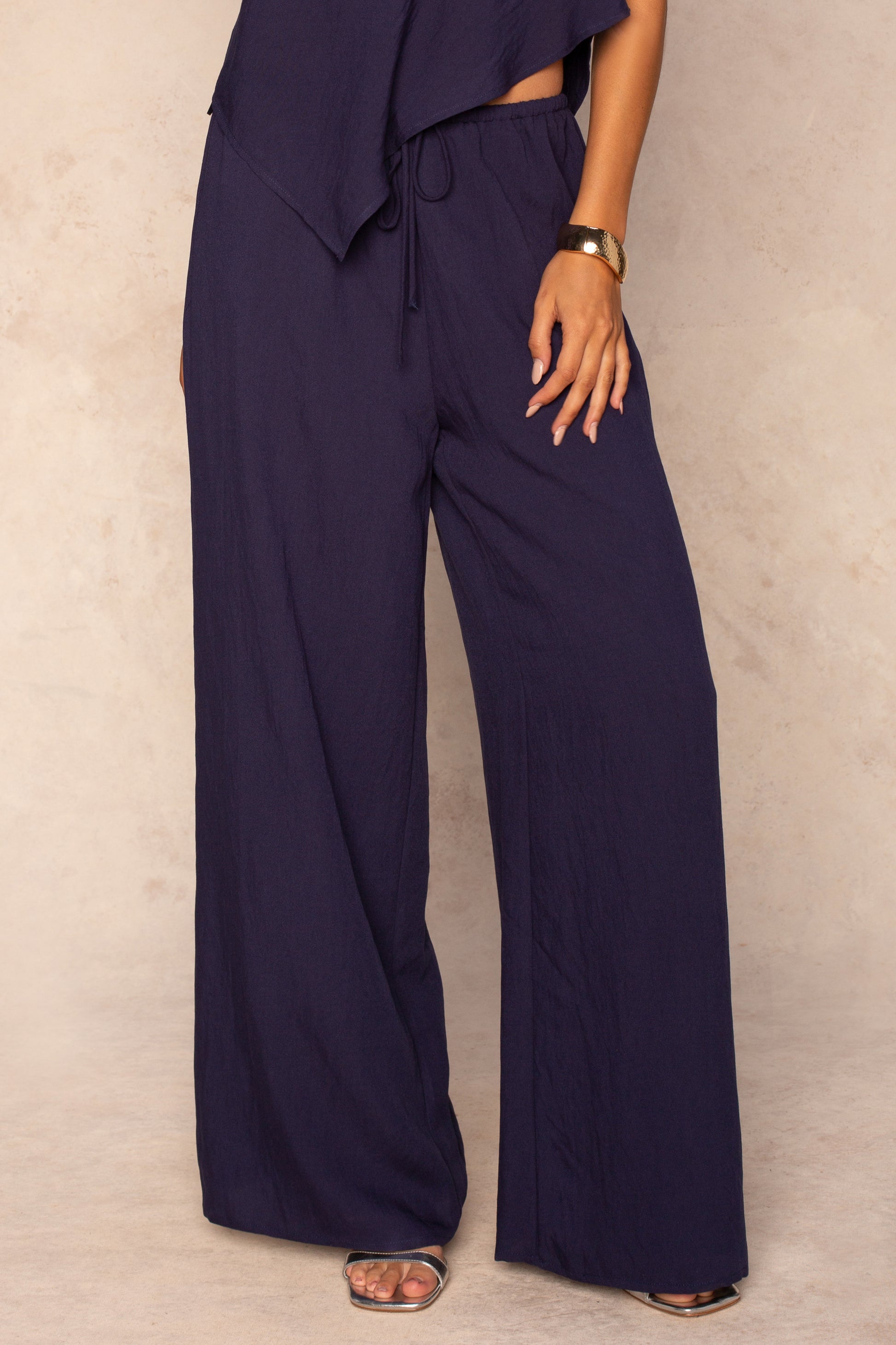 A person wears the Georgie Grayton Beach Pant in NAVY with an elastic waistband and matching top, styling it with a gold bracelet and open-toe sandals. The flowy fit pops against a beige background, one hand resting on their hip.