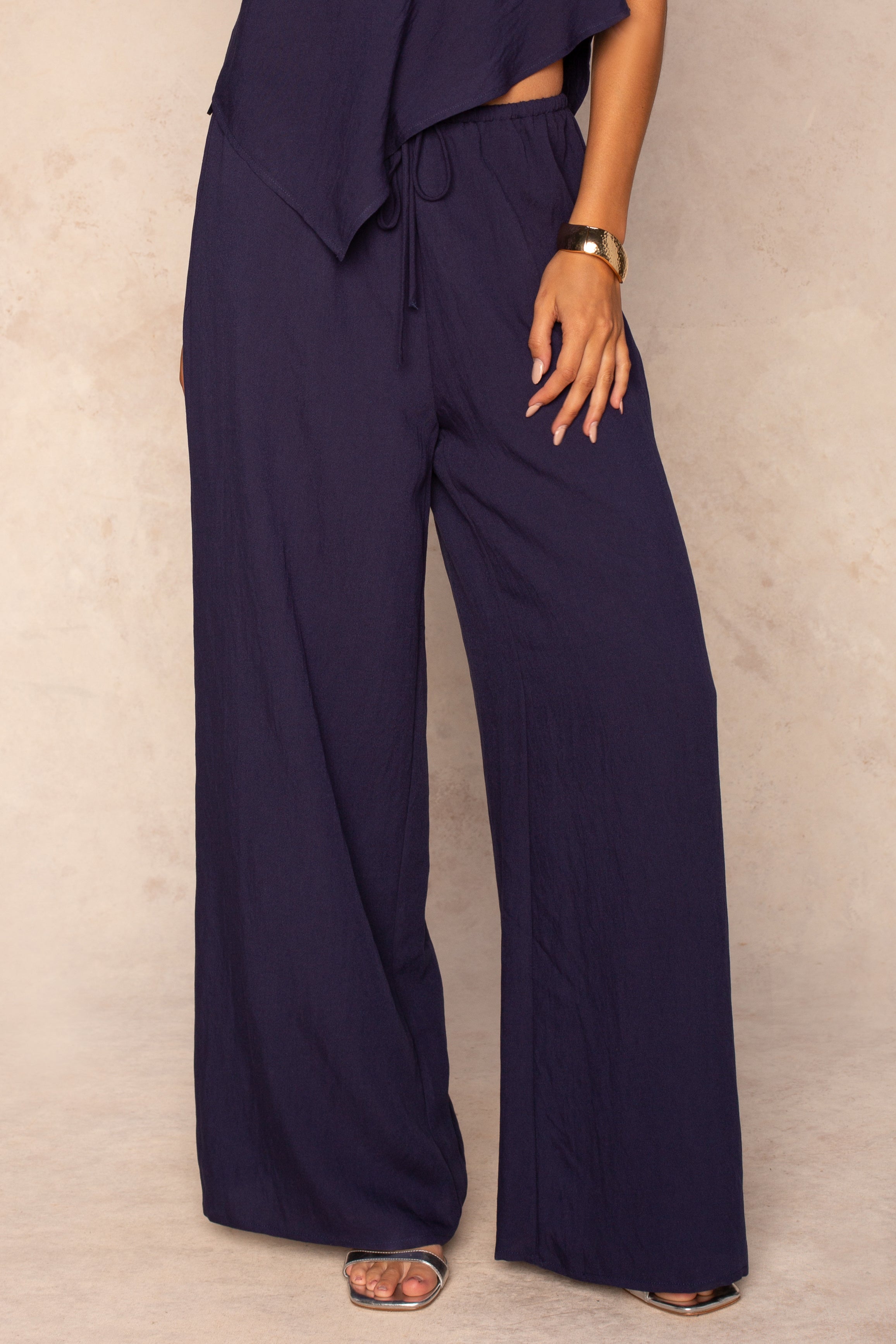 A person wears the Georgie Grayton Beach Pant in NAVY with an elastic waistband and matching top, styling it with a gold bracelet and open-toe sandals. The flowy fit pops against a beige background, one hand resting on their hip.