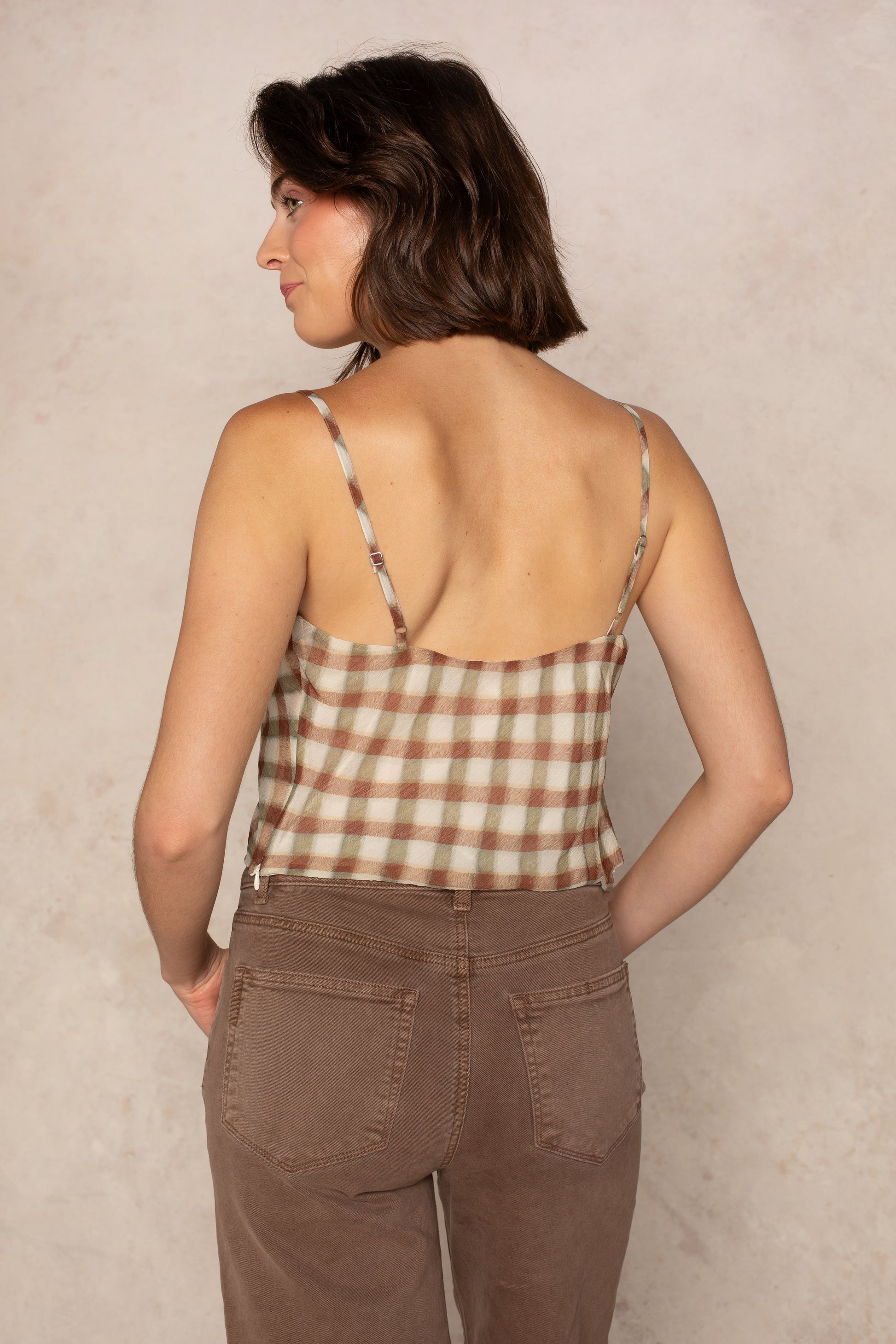 An Isla Naomi Chiffon Cami with adjustable spaghetti straps is worn by a woman with short brown hair, seen from behind. She pairs the plaid top with brown pants and stands against a neutral, light-colored background.