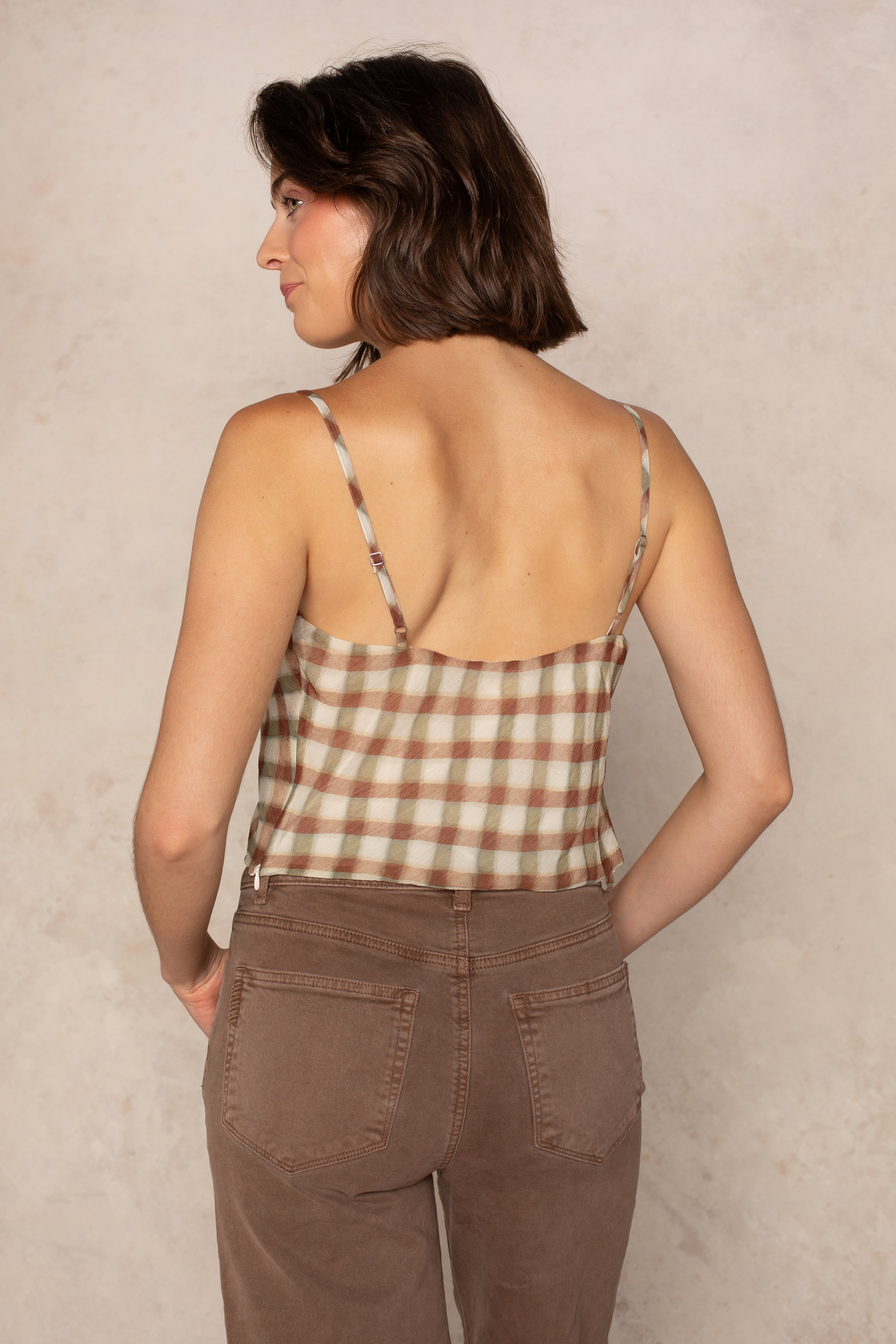 An Isla Naomi Chiffon Cami with adjustable spaghetti straps is worn by a woman with short brown hair, seen from behind. She pairs the plaid top with brown pants and stands against a neutral, light-colored background.
