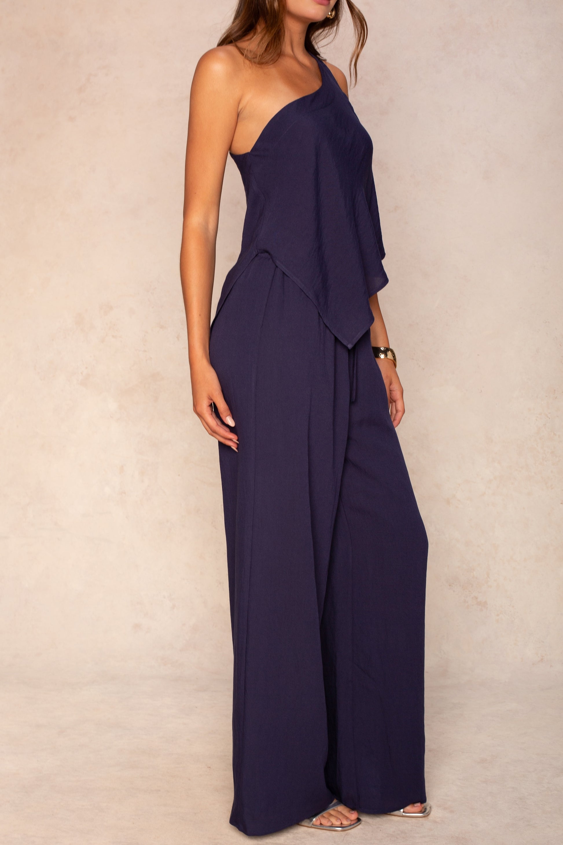 A woman wearing The Nora Set Top by Georgie in navy blue, featuring a one-shoulder design and draped silhouette, stands against a light beige textured background.