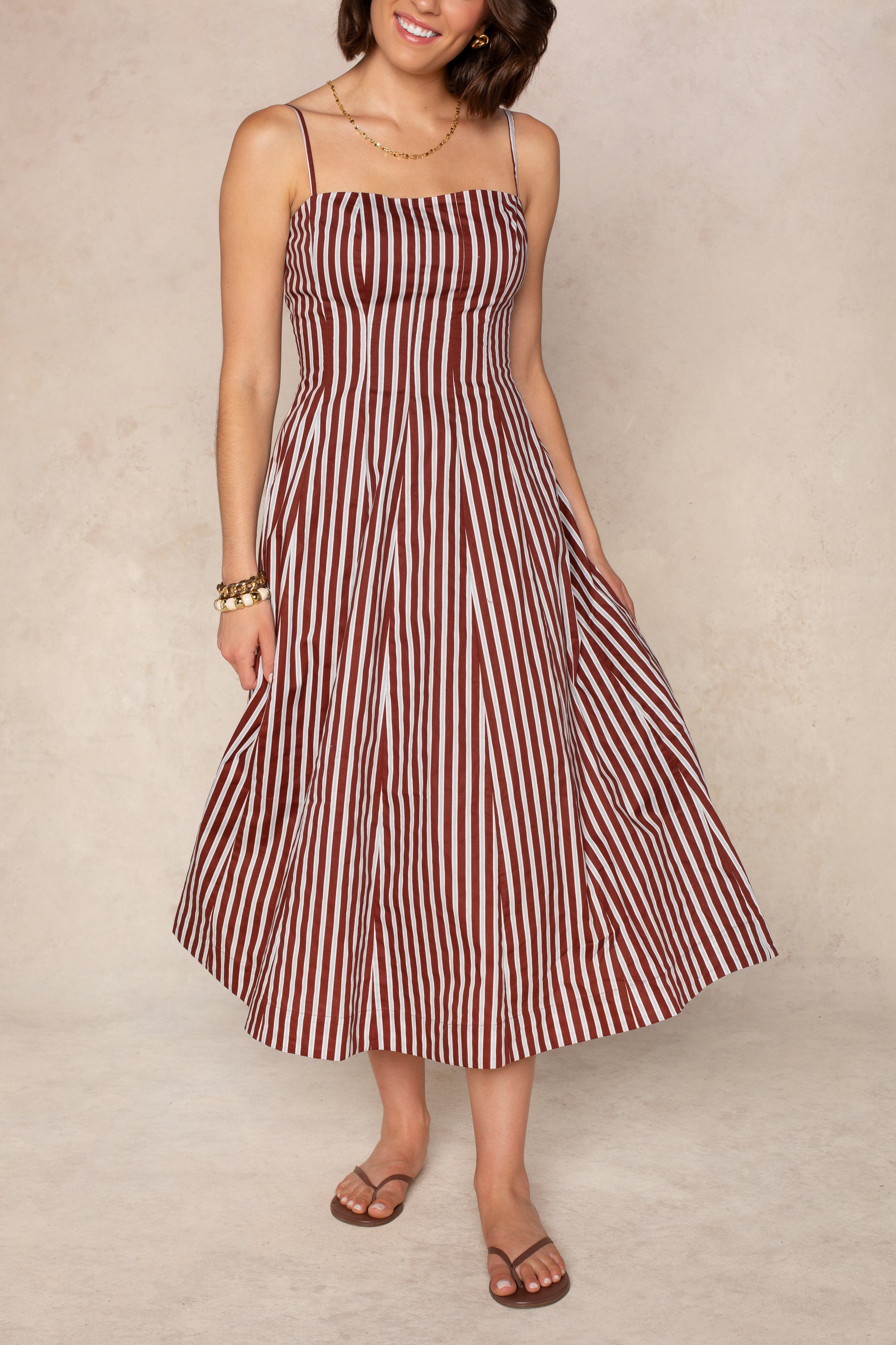 A woman stands against a neutral background wearing the Georgie Kenzie Striped Poplin Midi Dress, a sleeveless brown and white striped style with adjustable spaghetti straps. She completes the look with gold accessories and brown sandals.