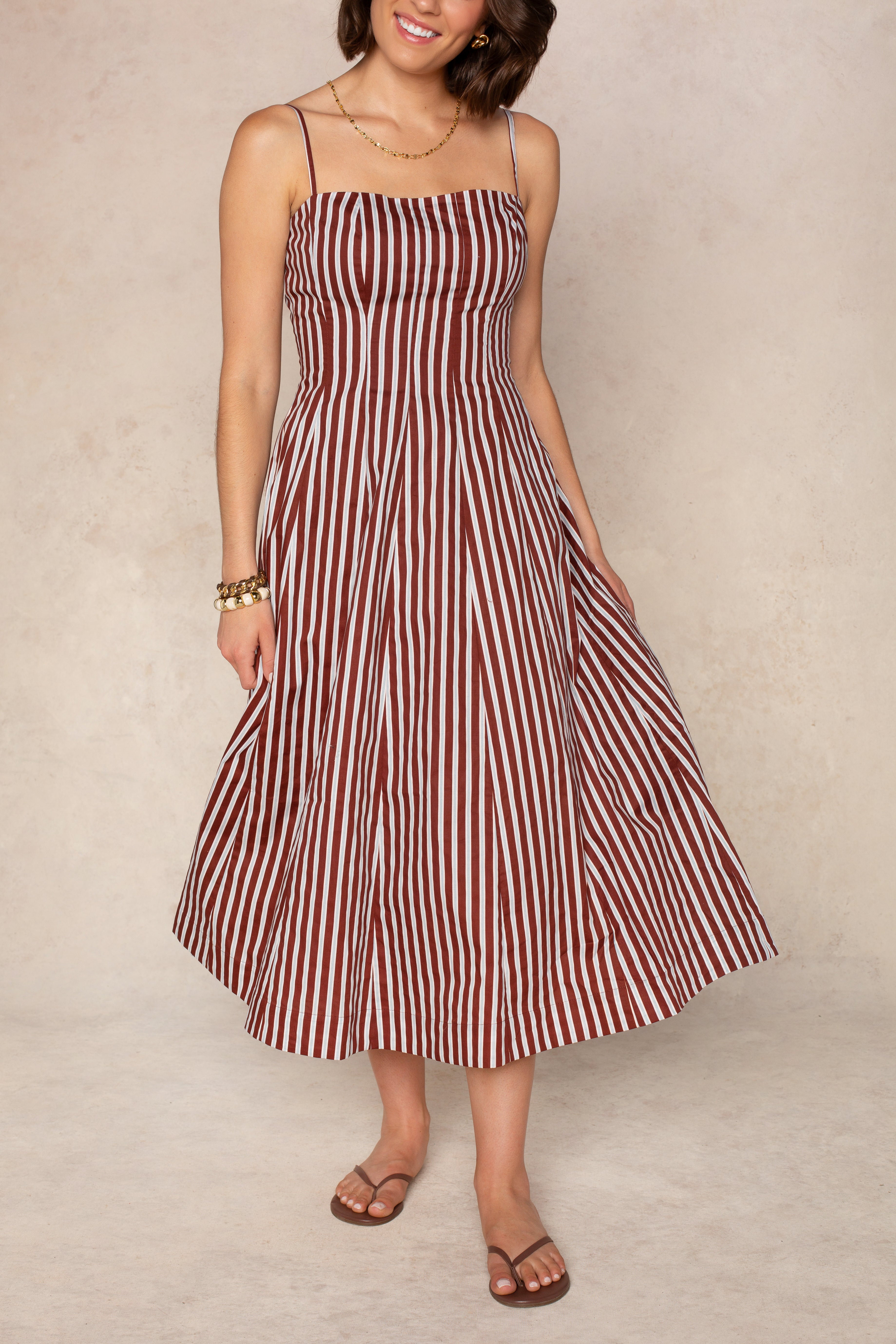 A woman stands against a neutral background wearing the Georgie Kenzie Striped Poplin Midi Dress, a sleeveless brown and white striped style with adjustable spaghetti straps. She completes the look with gold accessories and brown sandals.