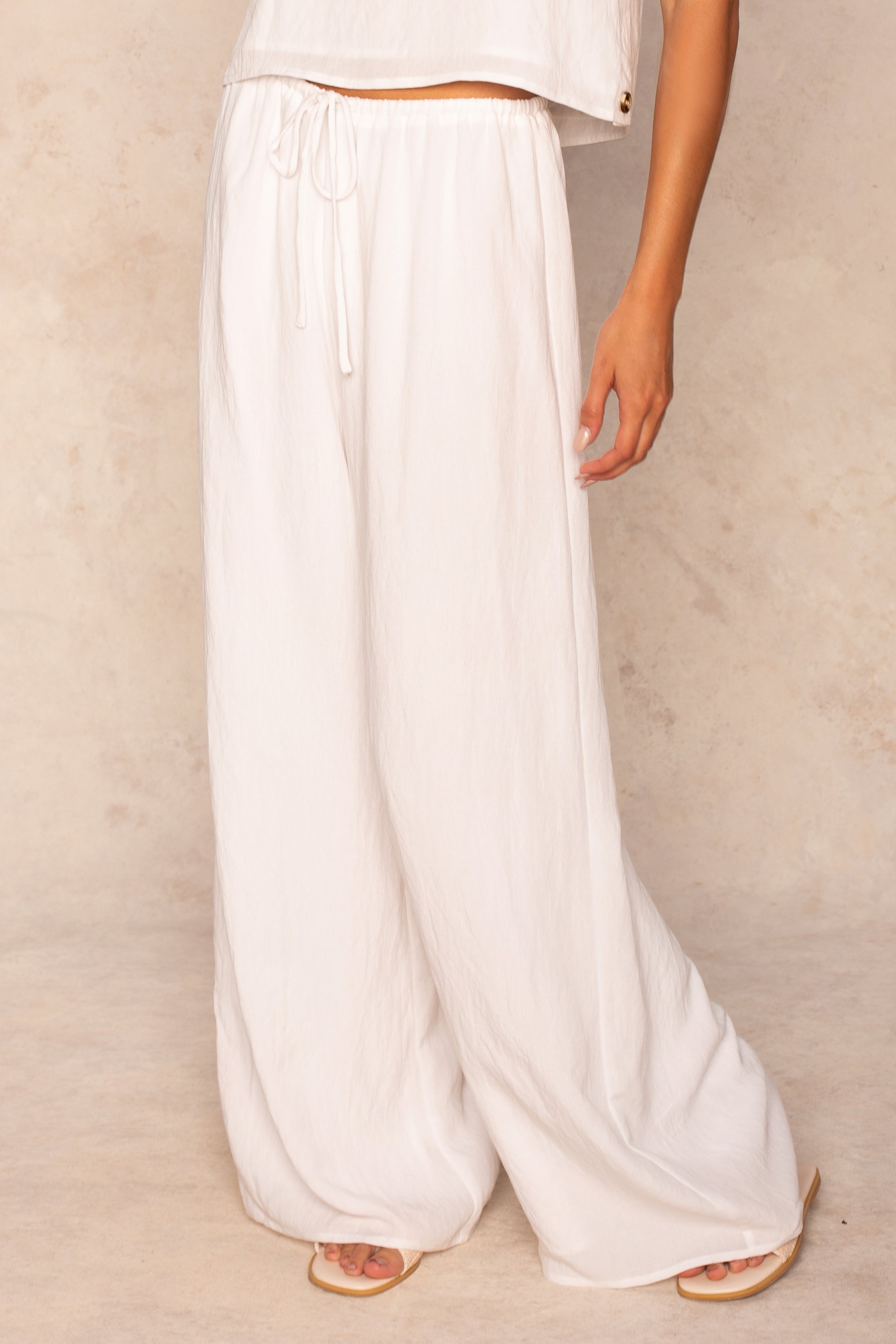 A person wears the Georgie Grayton Beach Pant in white with an elastic waistband and a matching sleeveless top, standing barefoot against a light beige background.