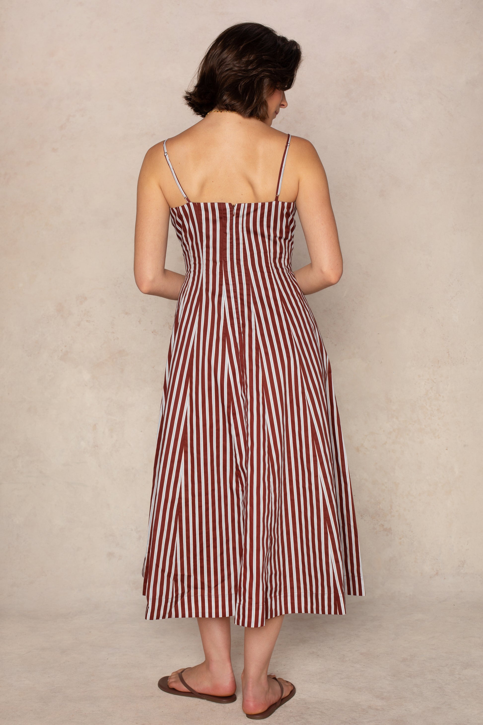 A woman with short brown hair wears the Georgie Kenzie Striped Poplin Midi Dress, a lightweight summer piece featuring vertical maroon and white stripes. The sleeveless, ankle-length dress drapes gracefully as she stands facing away from the camera, her arms relaxed by her sides. She completes the look with brown sandals.
