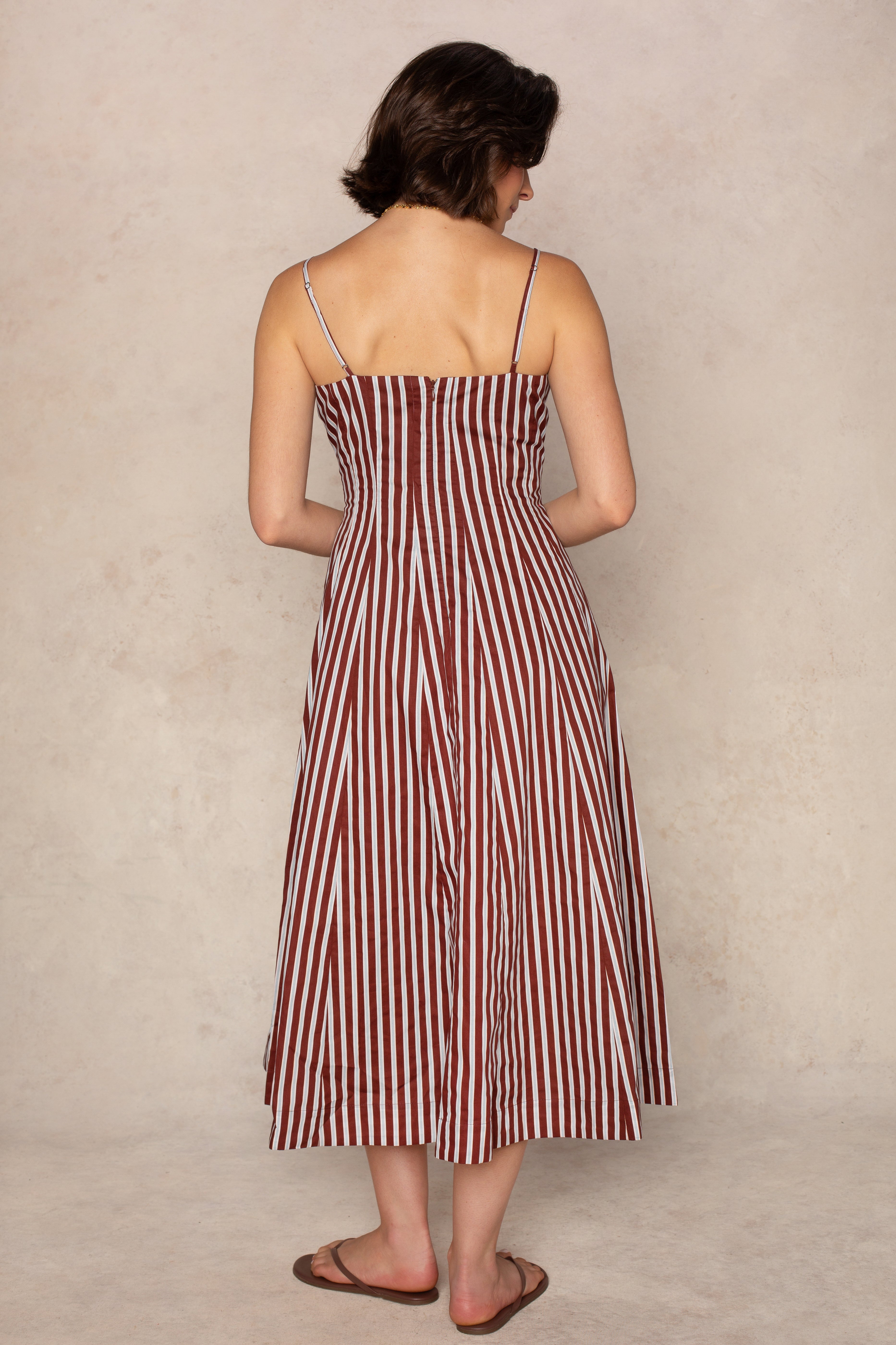 A woman with short brown hair wears the Georgie Kenzie Striped Poplin Midi Dress, a lightweight summer piece featuring vertical maroon and white stripes. The sleeveless, ankle-length dress drapes gracefully as she stands facing away from the camera, her arms relaxed by her sides. She completes the look with brown sandals.