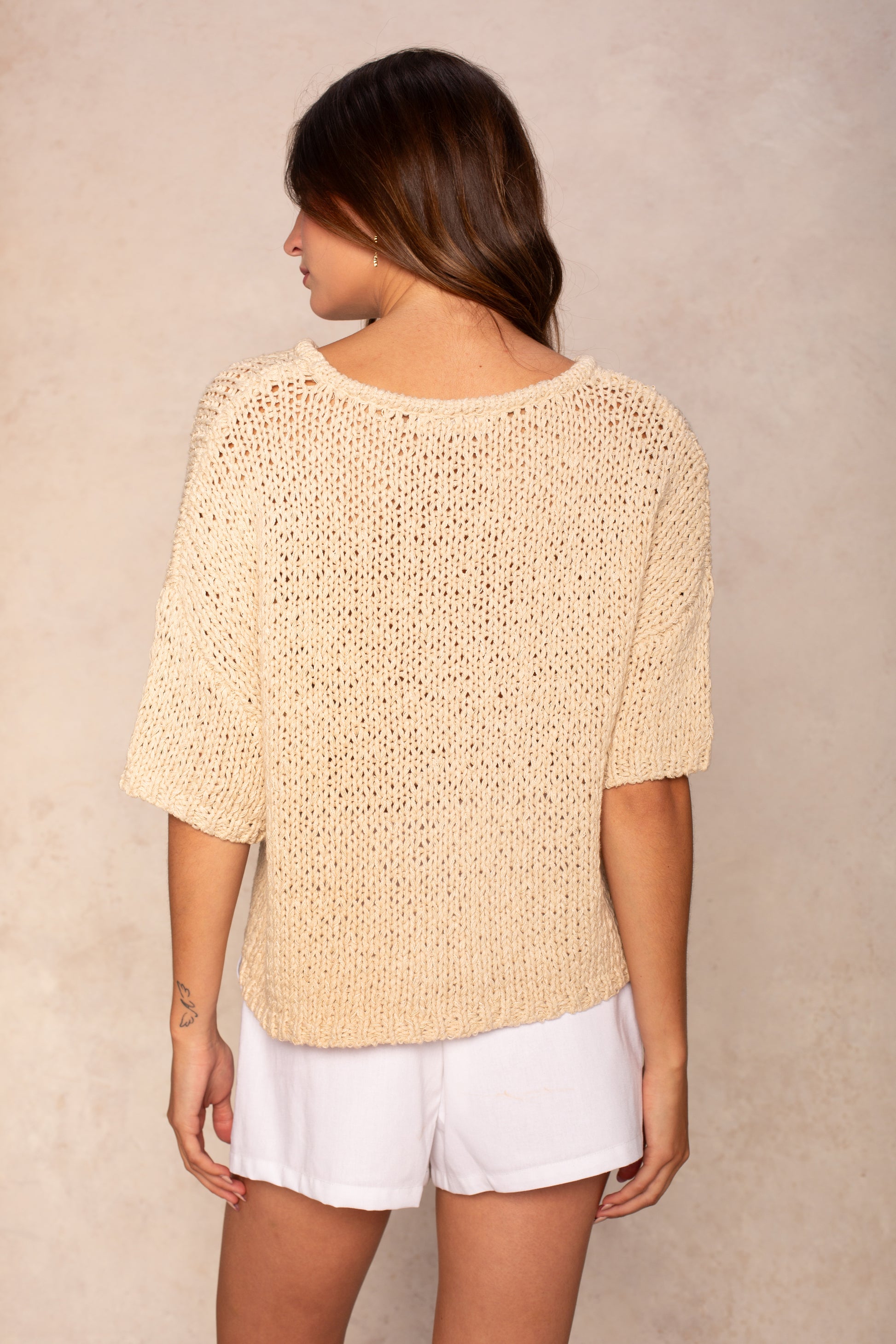 A woman with long brown hair stands with her back to the camera, wearing the Georgie Penny Short Sleeve Crew Neck Sweater in beige and white shorts, creating a relaxed look against a light, neutral background.