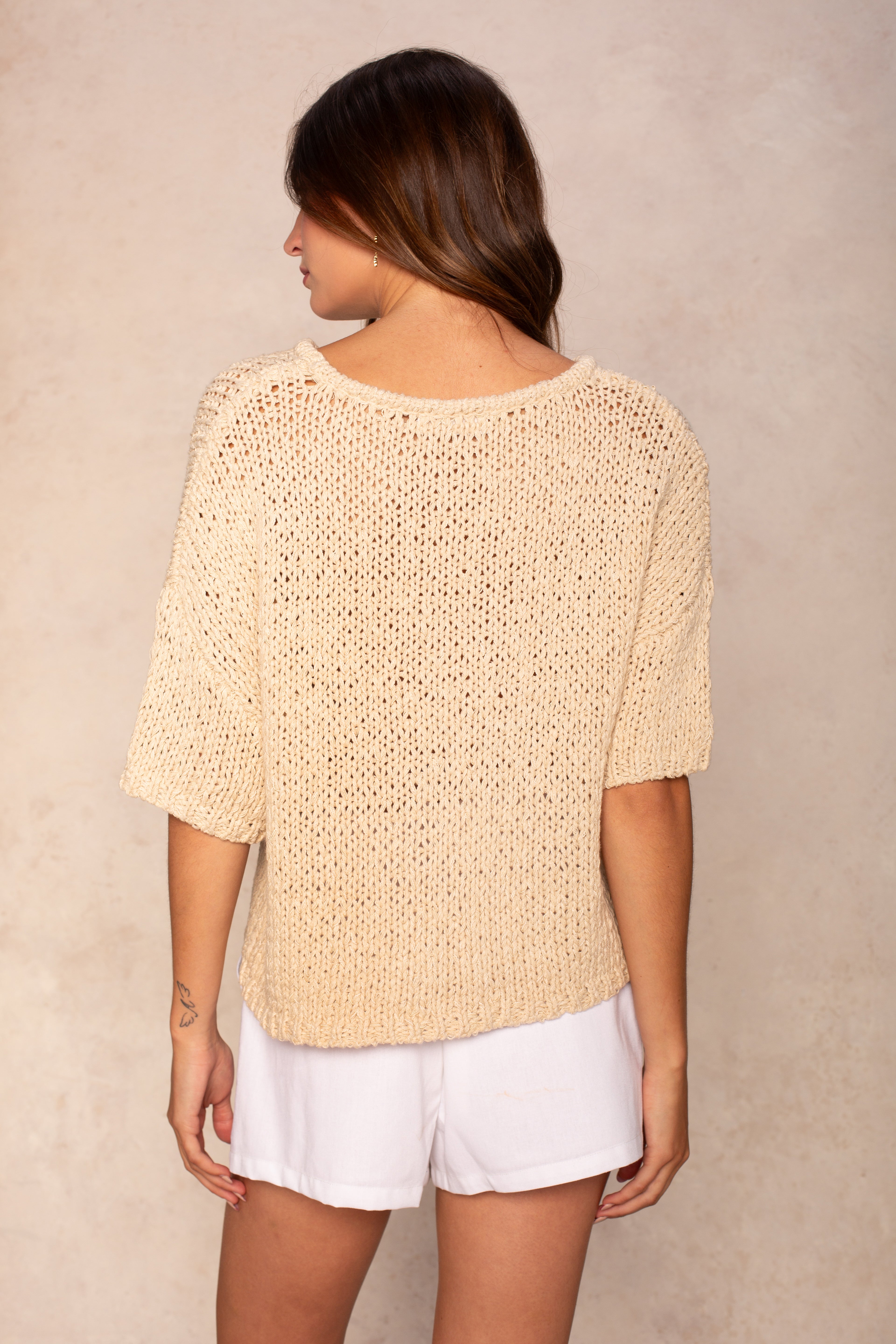 A woman with long brown hair stands with her back to the camera, wearing the Georgie Penny Short Sleeve Crew Neck Sweater in beige and white shorts, creating a relaxed look against a light, neutral background.