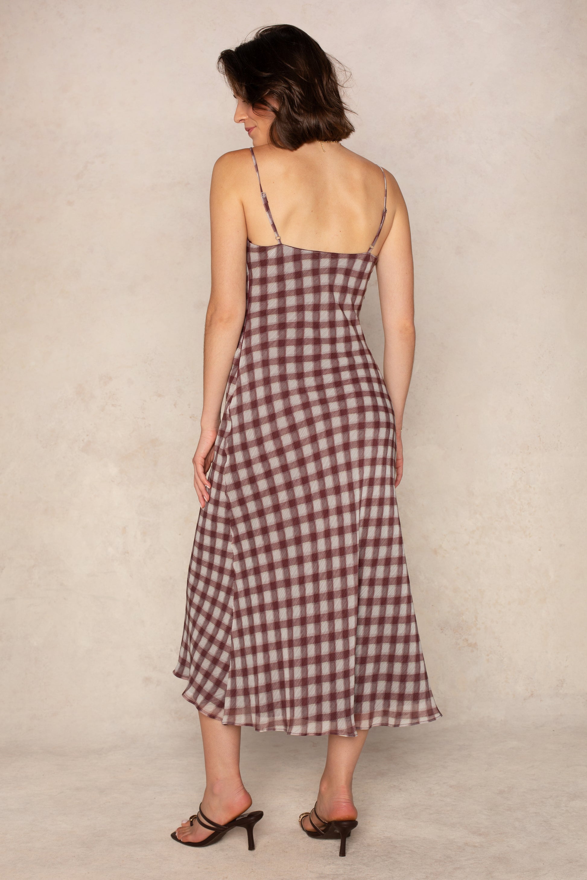 A woman with short brown hair models Isla's Pia Slip Maxi Dress, featuring a burgundy gingham pattern, bias-cut silhouette, and thin straps. She faces away in heeled sandals against a neutral textured background.