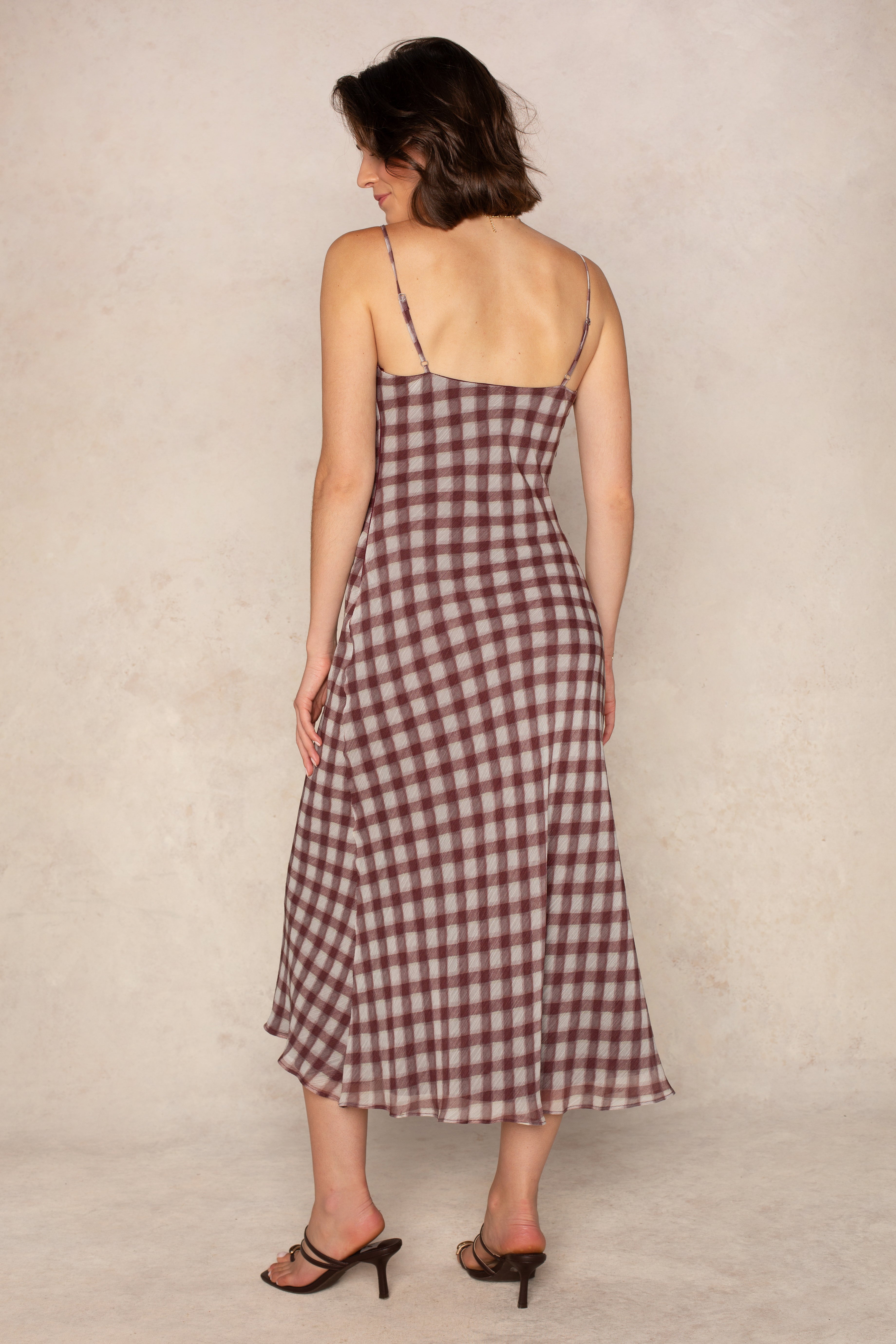 A woman with short brown hair models Isla's Pia Slip Maxi Dress, featuring a burgundy gingham pattern, bias-cut silhouette, and thin straps. She faces away in heeled sandals against a neutral textured background.