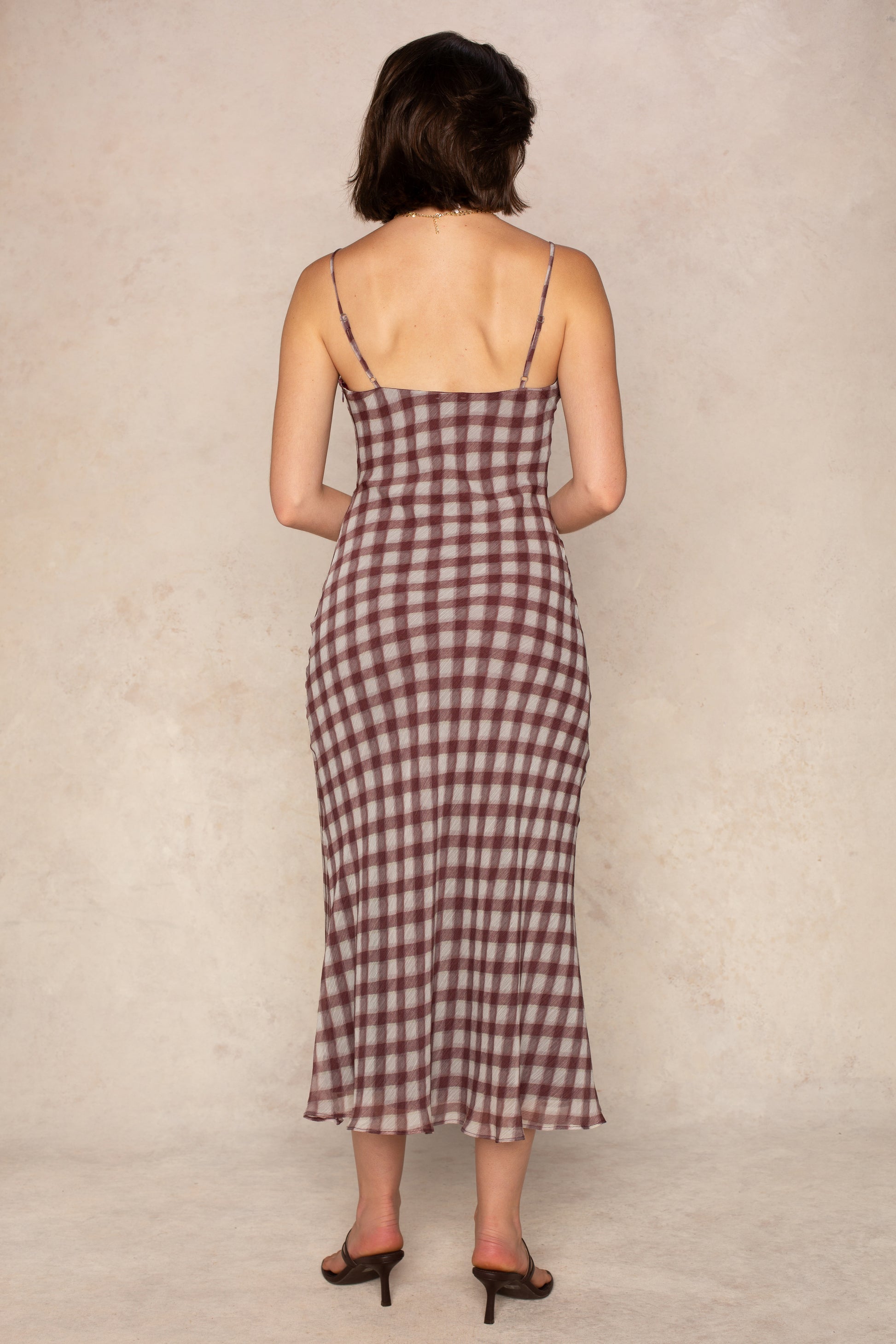 A woman faces away wearing the Isla Pia Slip Maxi Dress, a sleeveless burgundy gingham maxi with thin straps and a square neckline, paired with brown heeled sandals against a neutral beige background.
