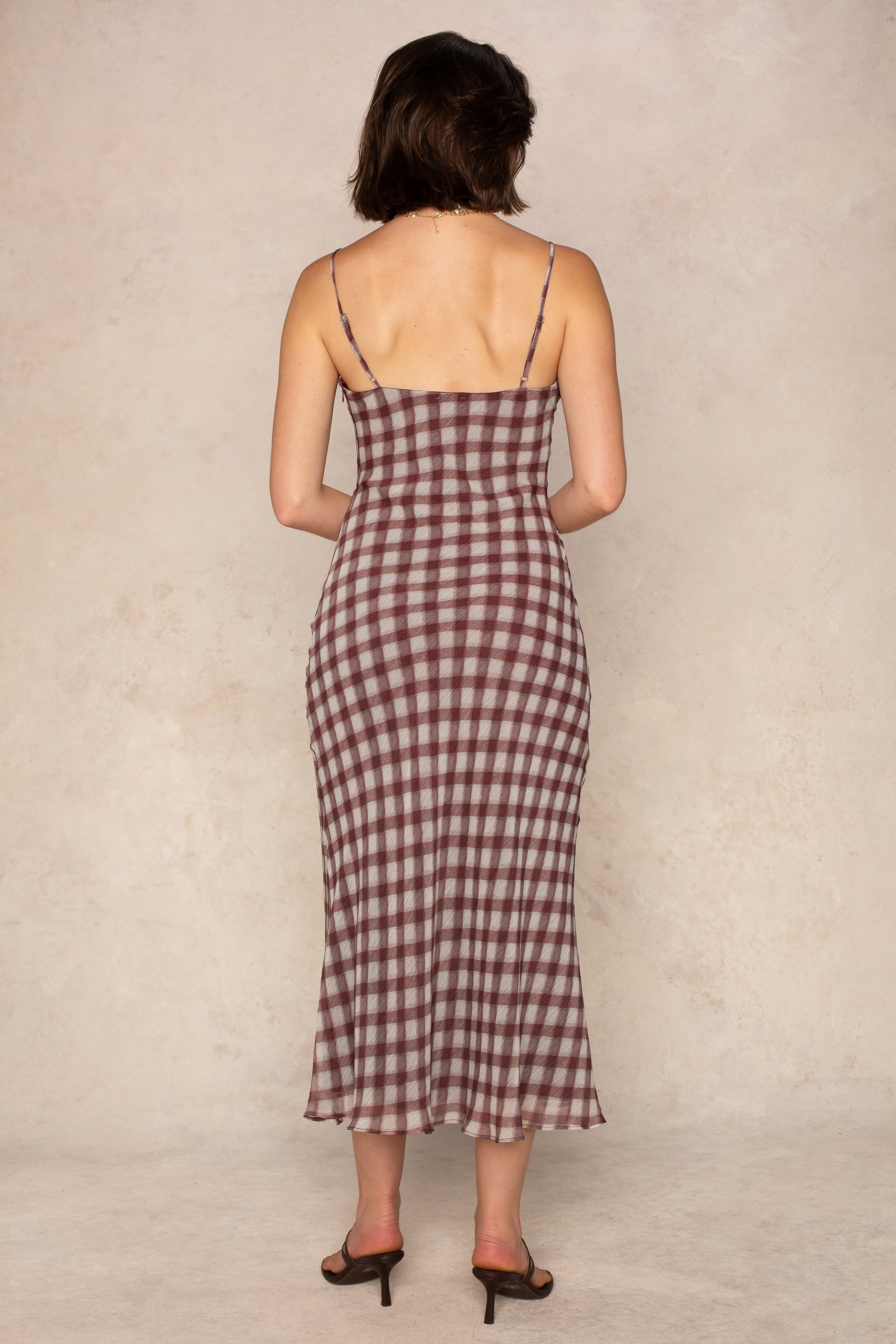 A woman faces away wearing the Isla Pia Slip Maxi Dress, a sleeveless burgundy gingham maxi with thin straps and a square neckline, paired with brown heeled sandals against a neutral beige background.